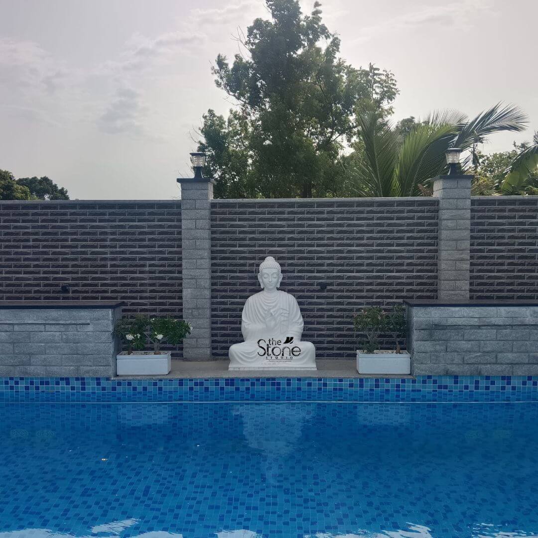 A serene eye-level photograph captures a luxurious outdoor swimming pool area featuring a magnificent 4-foot tall Fibre Buddha statue as the focal point. The statue rendered in a smooth matte white finish that mimics carved stone is seated in the traditional padmasana with hands in the dhyana mudra embodying profound tranquility and peace. It is positioned centrally on a raised stone platform directly at the water's edge appearing to watch over the crystal-clear blue tiled pool. The calm expression and detailed robes of the statue showcase the high craftsmanship of its lightweight fibre construction making it a substantial and durable piece of garden art. The background is dominated by a substantial boundary wall constructed from dark brown textured brick cladding offering a rich contrast to the white statue. This wall is punctuated by two light-gray granite pillars topped with modern globe-shaped outdoor lamps. To the left and right of the Buddha statue integrated into the same raised stone plinth are two rectangular white planters containing small manicured green shrubs adding a touch of nature and symmetry to the shrine-like setting. Above the wall lush green trees and a palm frond rise against a cloudy sky creating a private tropical oasis feel. The placement of this 4-foot Fibre Buddha statue transforms the swimming pool area into a tranquil retreat perfect for relaxation and meditation. The statue's size is impressive but not overwhelming creating a harmonious balance with the pool and the surrounding landscaping. Its elevated position at the water's edge makes it a stunning visual element visible from all parts of the pool deck and its light color stands out beautifully especially during the day. This image serves as an ideal inspiration for anyone looking to incorporate elegant Eastern-inspired elements into their swimming pool decor.