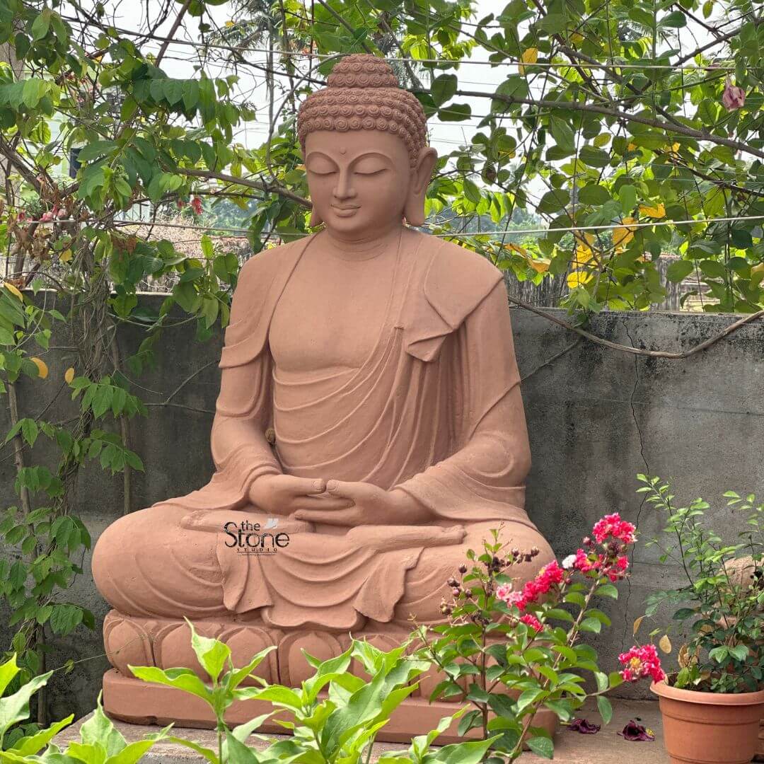 5 Feet Sandstone Buddha Statue for Garden 3 A magnificent 5-foot sandstone Buddha idol designed for a garden setting positioned amidst lush greenery and vibrant flowers as detailed in the accompanying article. The entire statue is carved from natural reddish-brown sandstone giving it an earthy warm and timeless appearance that harmonizes beautifully with its natural surroundings. The Buddha is depicted seated in the full lotus position with both hands gently resting in his lap palms facing upwards in the Dhyana Mudra symbolizing deep contemplation and inner peace. The robes are elegantly draped over his left shoulder with realistic flowing folds and pleats meticulously carved into the sandstone conveying a sense of serenity and soft fabric. The Buddha's face is characterized by a profoundly calm and contemplative expression with downcast half-closed eyes a gentle nose and subtle lips embodying a state of complete enlightenment and inner stillness. His tightly curled hair a distinctive iconographic feature is intricately textured and his elongated earlobes are visible symbolizing wisdom and a royal past. The Buddha is seated on a beautifully carved lotus pedestal also made of sandstone with individual lotus petals clearly defined symbolizing purity and spiritual awakening. The statue is surrounded by various potted plants and flowering bushes including vibrant pink flowers in the foreground enhancing the natural and tranquil ambiance. A concrete wall covered with climbing vines forms the background further integrating the idol into a peaceful garden sanctuary. This 5-foot sandstone Buddha idol is a powerful representation of peace and reflection making it a perfect and durable addition to enhance the aesthetic and spiritual ambiance of any garden or outdoor space.