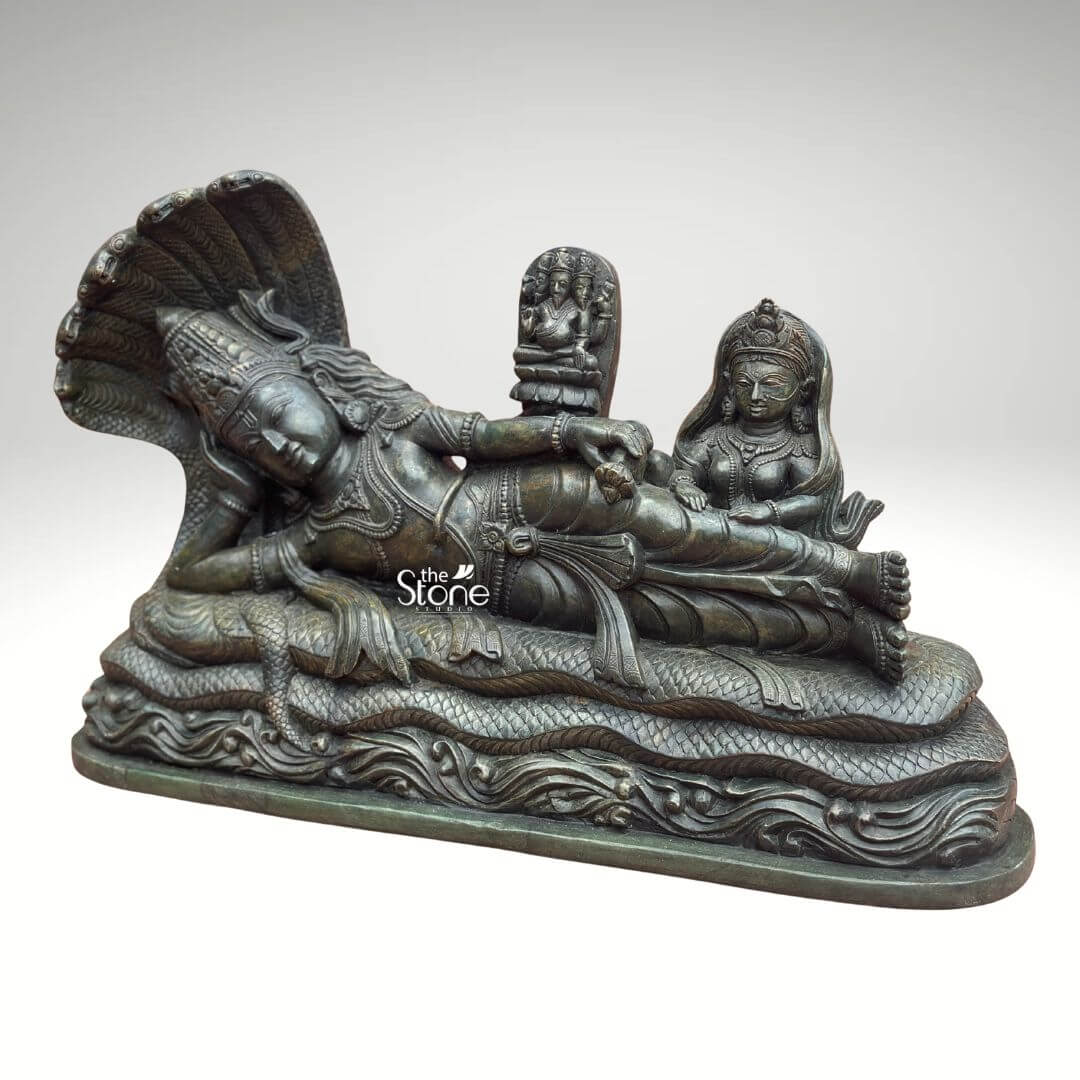 Ananta Shayana Vishnu Statue 20" 2 A striking green stone idol depicts Lord Vishnu in the majestic Ananta Shayana posture reclining on the coiled body of the multi-headed serpent Sheshnag. The intricate carving highlights the profound narrative of cosmic sleep and creation. The deep green hue of the stone possibly a type of serpentine or steatite imbues the sculpture with an ancient and sacred feel emphasizing the intricate details of the figures and the dynamic flow of the serpent's coils. Lord Vishnu is shown in a state of serene cosmic slumber his eyes gently closed symbolizing the period of universal dissolution. His right arm is bent supporting his head while his left arm rests along his side. He is elaborately adorned with a crown necklaces armlets and a dhoti all carved with meticulous precision. From his navel emerges a small lotus stem blossoming into a smaller depiction of Brahma the creator god signifying the cyclical emergence of creation from Vishnu's dream. At Vishnu's feet Goddess Lakshmi is seated attending to him with devotion completing the iconic scene of cosmic preservation. This green stone Ananta Shayana Vishnu idol is a profound artistic and spiritual representation of Hindu cosmology particularly Vishnu's role as the preserver of the universe. The choice of green stone often associated with growth and renewal enhances the idol's spiritual significance. This exquisitely detailed statue is an ideal piece for a grand temple a prominent home altar or a spiritual garden offering a powerful focal point for meditation and devotion and enriching any space with its deep cultural and religious resonance.