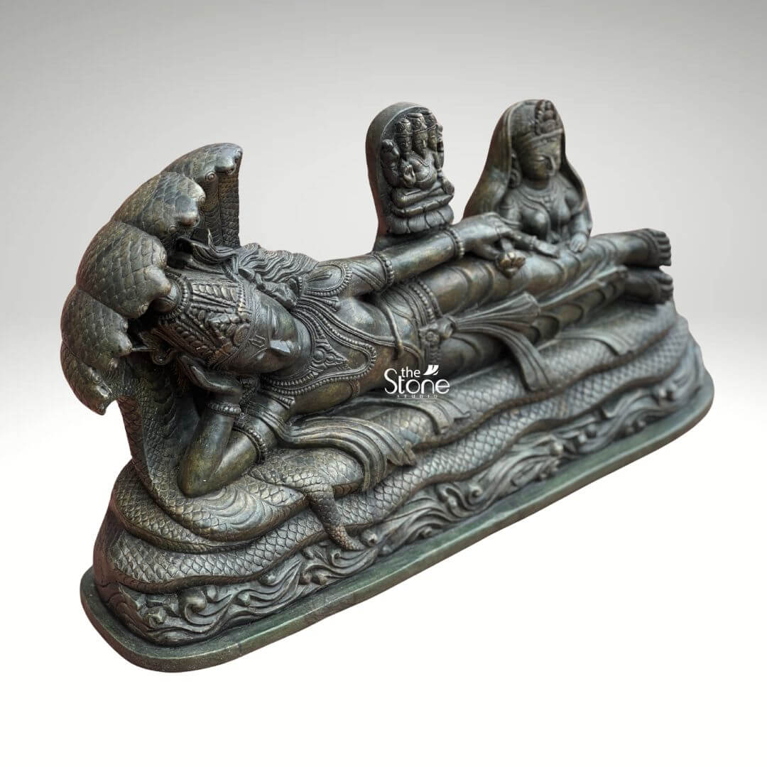 Ananta Shayana Vishnu Statue 20" 4 A magnificent green stone statue captures the iconic scene of Lord Vishnu in his Ananta Shayana postur reclining upon the cosmic serpent Sheshnag. The detailed carving viewed from a slightly elevated angle reveals the profound narrative of creation and cosmic repose. The statue's rich deep green stone possibly soapstone or a similar mineral lends a timeless and majestic quality to the intricate figures emphasizing the spiritual depth of this ancient Hindu motif. Lord Vishnu is depicted in a state of serene cosmic sleep his head gently supported by his right arm and his face bearing an expression of ultimate tranquility. His body is adorned with finely carved traditional jewelry including a crown necklaces armlets and a sacred dhoti all rendered with impressive precision. The multi-headed Sheshnag coils beneath him its numerous hoods fanning out to form a protective canopy over Vishnu's head. From Vishnu's navel a small lotus rises bearing a miniature figure of Brahma the creator god symbolizing the universe's cyclical creation from Vishnu's dream. To Vishnu's right seated attentively is Goddess Lakshmi his divine consort her form equally adorned and sculpted with reverence. This green stone Ananta Shayana Vishnu statue is a powerful and sacred representation of preservation cosmic order and the cyclical nature of time in Hindu mythology. The choice of green stone often associated with natural harmony and spiritual renewal enhances the idol's profound symbolism. This exquisitely detailed statue is a fitting centerpiece for a grand temple a significant home altar or a spiritual garden offering a rich source of contemplation and devotion and bringing an aura of ancient wisdom and peace to its surroundings.