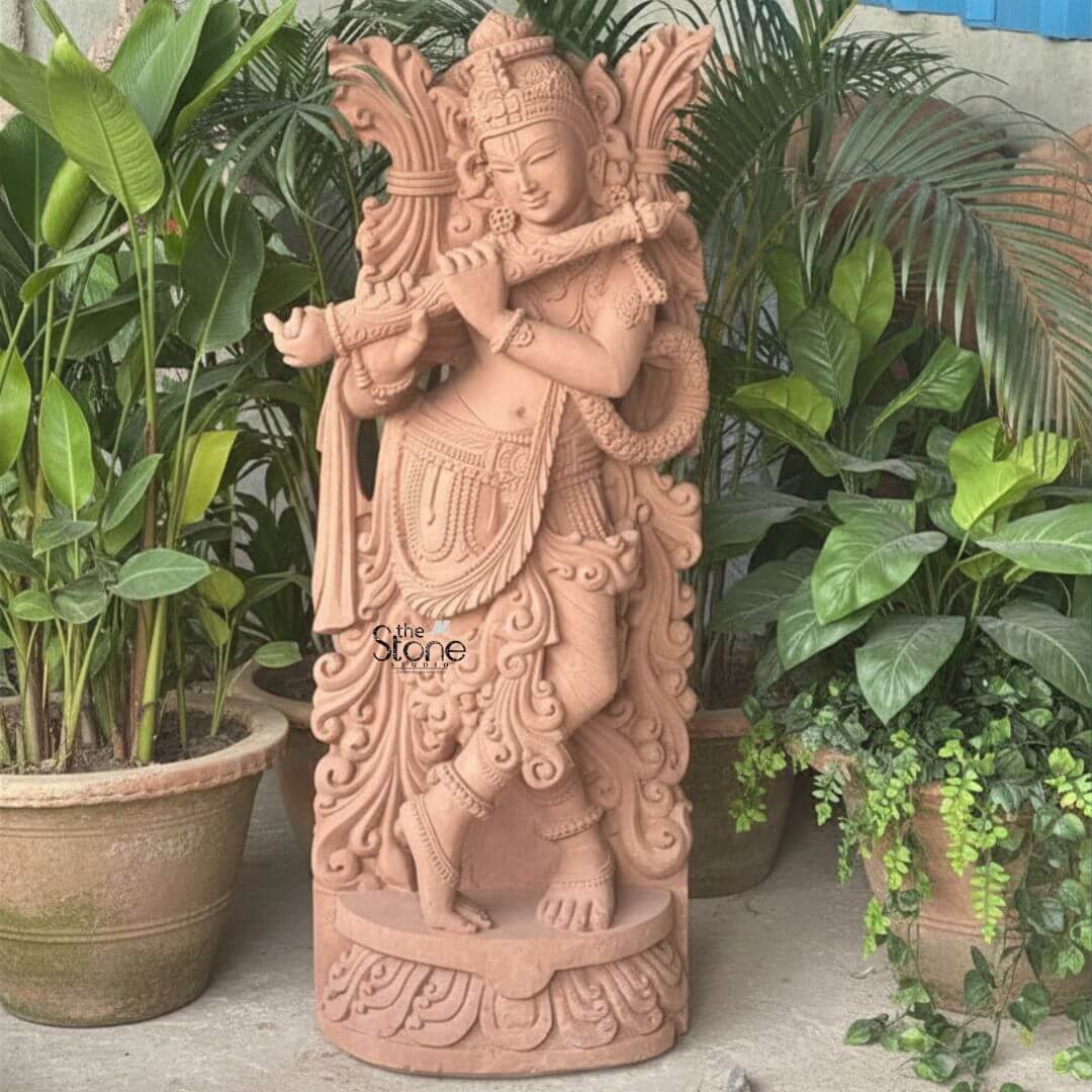 Lord Krishna Murti with Flute 3.5ft 3 A captivating 3.5-foot antique finish Krishna flute statue meticulously carved from sandstone displayed outdoors amidst lush green plants as featured in an article about such sculptures. The entire statue rendered in the natural reddish-brown hue of sandstone possesses an aged rustic charm consistent with an antique finish. Lord Krishna is shown standing in his iconic Tribhanga pose with one leg crossed gracefully in front of the other exuding divine playfulness and charm. He holds a flute to his lips with both hands appearing to play an enchanting melody. The carving details on Krishna's attire including his dhoti sash and intricate jewelry such as necklaces armlets, and ankle bracelets are exceptionally refined. He wears an elaborate crown and a garland or Vanamala drapes diagonally across his body. The flowing drapery of his garments is skillfully sculpted adding a sense of movement and grace to the figure. Behind Krishna an intricately carved design possibly representing foliage or an artistic prabhavali further enhances the visual richness and antique appeal of the murti. The sculpture stands on a circular ornate pedestal also crafted from sandstone featuring traditional Indian architectural motifs and decorative patterns adding to its grandeur and stability. The texture of the sandstone is visible throughout providing depth and authenticity to the artwork. This 3.5-foot sandstone Krishna statue is a powerful representation of devotion and artistic mastery perfectly suited for enhancing the aesthetic and spiritual ambiance of any home or garden space with its antique finish as discussed in the accompanying article.