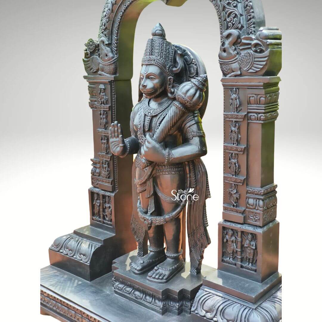 Standing Bajrangbali Statue 4ft 10 An impressive black stone Hanuman statue depicting Bajrangbali stands within an intricately carved prabhavali presented against a plain light background. This stunning black stone statue captures Lord Hanuman in a powerful and dynamic standing posture exuding an aura of immense strength devotion and courage. The dark polished surface of the stone provides a dramatic contrast enhancing the intricate details of the sculpture and its elaborate architectural frame making it a profound focal point for worship. Lord Hanuman is depicted with his characteristic features including a majestic crown prominent ear ornaments and a strong muscular physique. His right hand is raised in the Abhaya Mudra offering blessings and protection while his left hand holds his Gada symbolizing his immense power and readiness to vanquish evil. A garland of beads adorns his neck and a decorative sash drapes across his body. He stands on a lotus pedestal which is integrated into a multi-tiered base adorned with smaller figures and decorative carvings all contributing to the grandeur of the piece. The intricate prabhavali above and behind him is equally detailed featuring divine figures floral motifs and a Kirtimukha at the apex flanked by mythical creatures. This black stone Bajrangbali statue with its prabhavali is a profound representation of Lord Hanuman the epitome of strength devotion and selfless service. The exceptional craftsmanship in black stone not only makes it a visually striking piece but also embodies deep cultural and religious significance offering a powerful focal point for devotion and prayer. The rich detailing of the deity and his surrounding archway reflects centuries of tradition making it a truly remarkable and revered artifact that brings blessings protection and a spiritual presence into any sacred space.