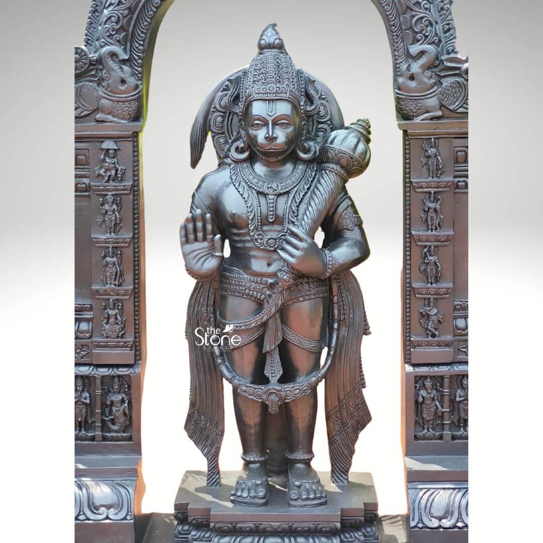 Standing Bajrangbali Statue 4ft 4 An impressive black granite Hanuman statue framed by an intricately carved temple archway stands as a powerful and revered figure against a plain light background making it an ideal piece for a home. This stunning black stone statue captures Lord Hanuman in a dynamic standing posture exuding an aura of immense strength devotion and courage. The dark polished surface of the black granite provides a dramatic contrast enhancing the intricate details of the sculpture and its elaborate architectural frame creating a striking focal point for any sacred space. Lord Hanuman is depicted with his characteristic features including a majestic crown prominent ear ornaments and a strong muscular physique that symbolizes his extraordinary power. His right hand is raised in the Abhaya Mudra offering blessings and protection to devotees while his left hand holds his Gada symbolizing his immense power and readiness to vanquish evil. A garland of beads adorns his neck and a decorative sash drapes across his body all carved with meticulous precision. He stands on a lotus pedestal which is integrated into a multi-tiered base adorned with smaller figures and decorative carvings all contributing to the grandeur of the piece. The intricate temple archway above him is equally detailed featuring divine figures floral motifs and a Kirtimukha at the apex flanked by mythical creatures. This black granite Hanuman statue is a profound representation of Lord Hanuman the epitome of strength devotion and selfless service. The exceptional craftsmanship in black granite not only makes it a visually striking piece but also embodies deep cultural and religious significance offering a powerful focal point for devotion and prayer in a home. The rich detailing of the deity and his surrounding archway reflects centuries of tradition making it a truly remarkable and revered artifact that brings blessings protection and a spiritual presence into any household.