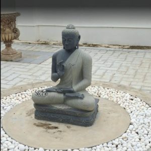 A serene 3 feet Black Stone Buddha Statue meticulously carved from blackstone with subtle grey undertones sits peacefully in a modern outdoor garden setting possibly in Kollam Kerala as suggested by its article context. This Black Stone Buddha Statue in Kollam Kerala depicts the Buddha in a meditative posture seated cross-legged on a lotus base. His right hand is raised in the Vitarka Mudra signifying teaching or intellectual discussion while his left hand rests in his lap. His expression is one of profound tranquility and wisdom with downcast eyes embodying deep spiritual introspection. The statue features a distinctive two-toned appearance: the head and upper body are of a darker rich blackstone while the draped robes are a lighter grey creating a beautiful contrast that highlights the figure's form. His hair is styled in tightly coiled curls and the is subtly visible. The flowing drapery of his robes is finely sculpted, showing soft folds and creases. The statue rests on a carved lotus-petal base which in turn sits within a circular concrete or stone area. Surrounding this circular area is a bed of pristine white pebbles or river stones creating a clean and minimalist aesthetic that enhances the statue's calming presence. In the background a patterned stone pavement extends along with a classical-style ornate stone planter suggesting a well-designed garden or courtyard. A plain white wall in the distance further emphasizes the tranquility of the setting. This detailed description effectively connects the visual elements of the 3 feet blackstone Buddha statue with its spiritual significance and aesthetic qualities, making it an ideal representation for an article about a Black Stone Buddha Statue in Kollam Kerala.