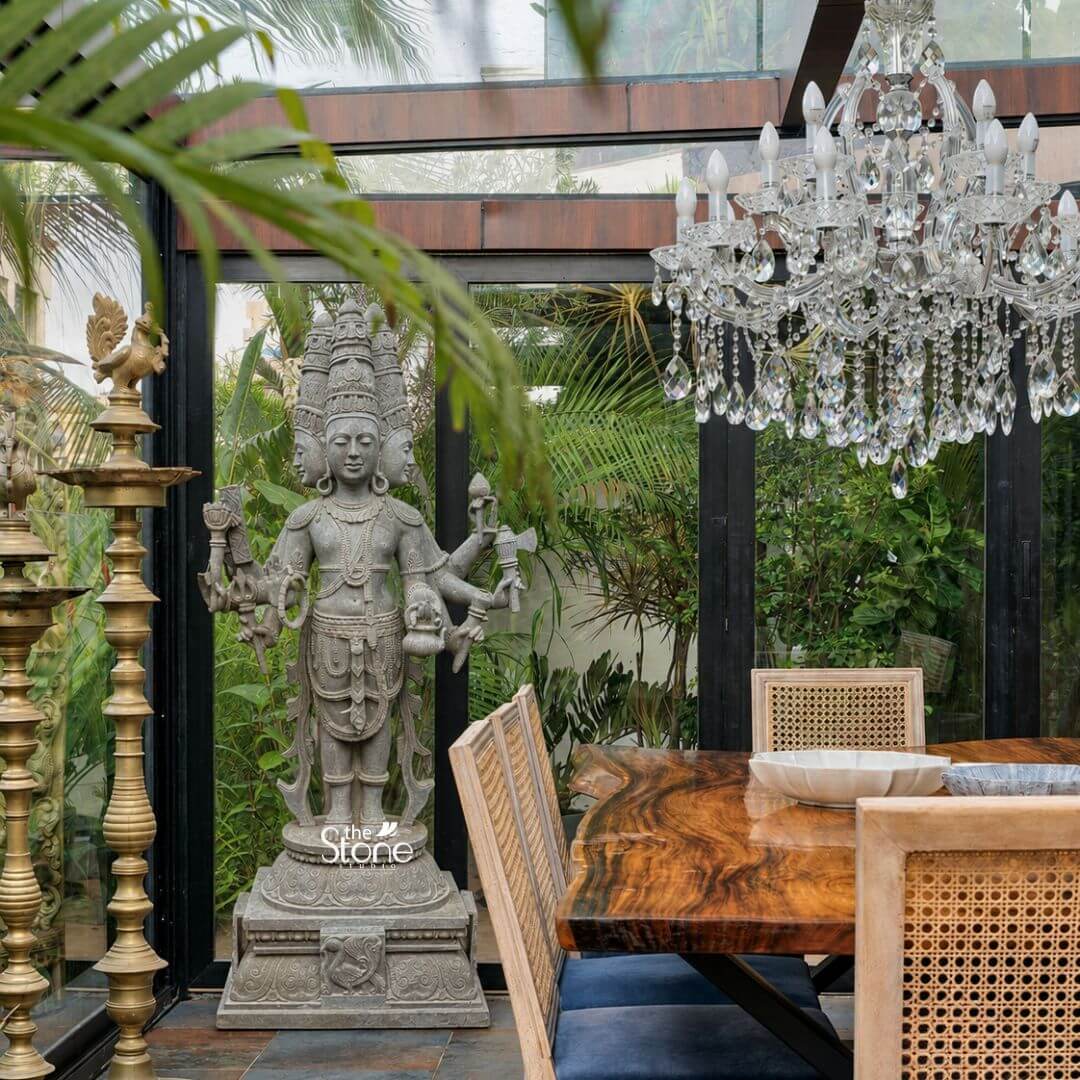 Brahma Stone Statue 7ft 3 A magnificent 7-foot greystone Brahma Dev Stone Statue is majestically displayed in a bright modern indoor space with large windows overlooking a lush garden creating a harmonious blend of spiritual art and contemporary design. The statue depicts Lord Brahma the creator god in a standing posture with his characteristic four heads symbolizing his omnipresence and knowledge. The natural grey tones of the stone lend an ancient and dignified quality to this impressive statue highlighting its detailed carvings amidst a sophisticated interior. Lord Brahma is meticulously carved with his four serene and contemplative faces facing the cardinal directions. He is richly adorned with an elaborate crown multiple necklaces armlets and a sacred thread all finely detailed. In his multiple hands he holds his traditional attributes including a sruva or rosary a kamandalu and a puskara symbolizing his role in creation and knowledge. His attire is elegantly draped and he stands barefoot on a lotus pedestal which in turn rests on a multi-tiered intricately carved base featuring smaller decorative elements. Adjacent to the statue towering brass oil lamps add to the traditional aesthetic while a modern wooden dining table with cane-back chairs and a crystal chandelier complete the elegant setting. This 7-foot greystone Brahma Dev Stone Statue is a profound representation of creation wisdom and cosmic knowledge serving as a powerful and auspicious addition to this home. The exceptional craftsmanship in natural greystone not only makes it a visually striking piece but also imbues it with deep cultural and religious significance. This exquisitely detailed statue integrated into a well-designed indoor-outdoor space serves as a commanding focal point radiating positive energy and ancient wisdom and fostering an environment of cultural richness and spiritual contemplation.