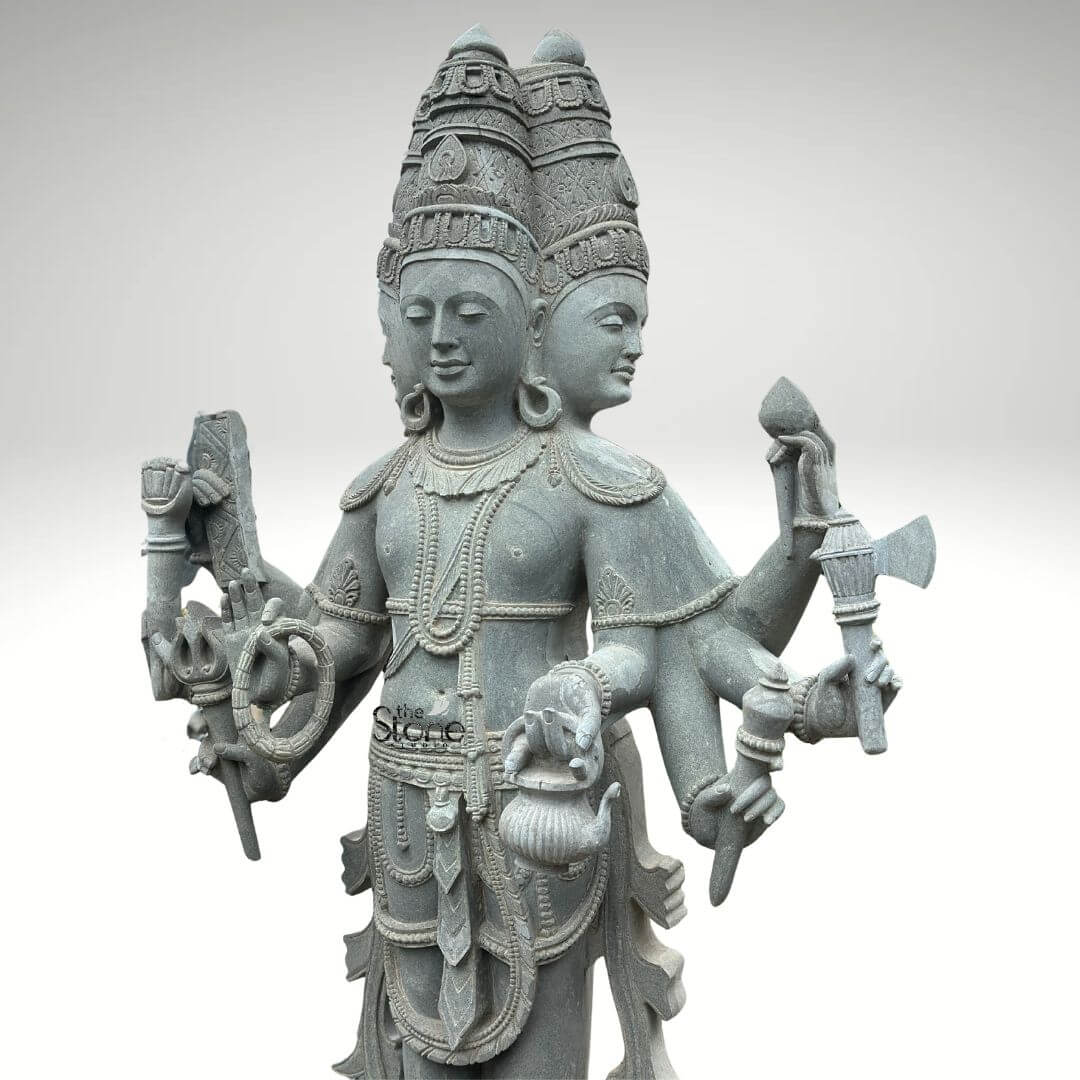 Brahma Stone Statue 7ft 4 A magnificent 7-foot greystone Brahma statue designed for temple installation is presented in a stately front-facing view against a plain light background emphasizing its grandeur and intricate craftsmanship. The statue depicts Lord Brahma the creator god in a standing posture with his characteristic four heads symbolizing his omnipresence and knowledge. The natural grey tones and subtle textures of the stone lend an ancient and dignified quality to this impressive statue highlighting its detailed carvings. Lord Brahma is meticulously carved with his four serene and contemplative faces each adorned with an elaborate crown and facing the cardinal directions. He is richly adorned with multiple necklaces armlets and a sacred thread all finely detailed. In his multiple hands he holds his traditional attributes including a sruva or rosary a kamandalu and a puskara symbolizing his role in creation and knowledge. His attire is elegantly draped and he stands barefoot on a lotus pedestal which in turn rests on a multi-tiered intricately carved base. The high level of detail in the carving from the delicate facial features to the intricate jewelry speaks to exceptional artistry. This 7-foot greystone Brahma statue is a profound representation of creation wisdom and cosmic knowledge making it a powerful and auspicious addition for temple installation. The exceptional craftsmanship in natural greystone not only makes it a visually striking piece but also imbues it with deep cultural and religious significance. This exquisitely detailed statue serves as a commanding focal point for devotion and prayer radiating positive energy and ancient wisdom and enriching any sacred temple space.