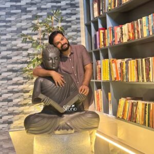 A warm and engaging image features a smiling man embracing a 3-foot Buddha fiber statue presented in a cozy indoor setting possibly a home or library in Kannur Kerala. The Buddha statue is depicted in a serene meditative pose with the man's arm gently draped around its shoulders highlighting the harmonious integration of spiritual art into personal space. The statue finished in a dark stone-like texture sits atop a light-colored cylindrical base contributing to a tranquil and intellectually rich atmosphere. The Buddha's face is sculpted with a calm and contemplative expression conveying deep inner peace. He is shown holding what appears to be a stringed musical instrument possibly a sitar or veena adding a unique and artistic dimension to the traditional Buddha iconography. The man with a beard and wearing a casual shirt smiles warmly at the camera leaning his head against the Buddha's. In the background a large bookshelf filled with numerous books creates a vibrant and intellectual backdrop while a textured gray wall and a potted plant add further depth to the scene. This 3-foot Buddha fiber statue is an exceptional piece that brings an immediate sense of calm and spiritual harmony to any environment as beautifully demonstrated by its appreciative owner. Its durable fiber construction ensures longevity while its artistic depiction combining meditation with music makes it a unique and inspiring presence. As a symbol of enlightenment and peace this Buddha statue is not only an artistic masterpiece but also a significant spiritual icon inviting contemplation and fostering a tranquil atmosphere within a welcoming home in Kannur Kerala.