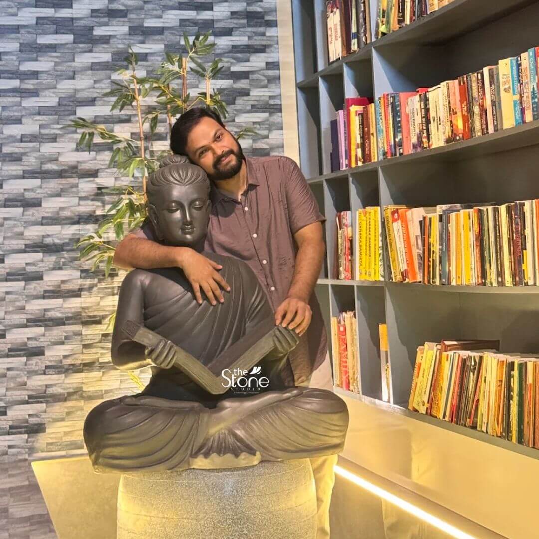 Buddha Statue With Book 3ft 3 A warm and engaging image features a smiling man embracing a 3-foot Buddha fiber statue presented in a cozy indoor setting possibly a home or library in Kannur Kerala. The Buddha statue is depicted in a serene meditative pose with the man's arm gently draped around its shoulders highlighting the harmonious integration of spiritual art into personal space. The statue finished in a dark stone-like texture sits atop a light-colored cylindrical base contributing to a tranquil and intellectually rich atmosphere. The Buddha's face is sculpted with a calm and contemplative expression conveying deep inner peace. He is shown holding what appears to be a stringed musical instrument possibly a sitar or veena adding a unique and artistic dimension to the traditional Buddha iconography. The man with a beard and wearing a casual shirt smiles warmly at the camera leaning his head against the Buddha's. In the background a large bookshelf filled with numerous books creates a vibrant and intellectual backdrop while a textured gray wall and a potted plant add further depth to the scene. This 3-foot Buddha fiber statue is an exceptional piece that brings an immediate sense of calm and spiritual harmony to any environment as beautifully demonstrated by its appreciative owner. Its durable fiber construction ensures longevity while its artistic depiction combining meditation with music makes it a unique and inspiring presence. As a symbol of enlightenment and peace this Buddha statue is not only an artistic masterpiece but also a significant spiritual icon inviting contemplation and fostering a tranquil atmosphere within a welcoming home in Kannur Kerala.