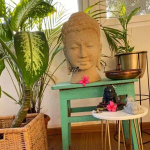 A serene 15-inch Buddha Head Statue carved from off-white sandstone is thoughtfully placed on a rustic green wooden stool becoming a focal point in a well-decorated home interior in Hyderabad. The Buddha head with its finely sculpted features embodies a sense of deep tranquility and spiritual wisdom. His eyes are gently closed in meditation with a subtle smile on his lips and elongated earlobes indicative of his princely past. The off-white sandstone material gives the Buddha head a natural earthy texture and a light calming presence that blends harmoniously with its surroundings. The hair is meticulously carved in tight curls culminating in the ushnisha the cranial bump symbolizing enlightenment. A single vibrant pink flower is delicately placed in front of the Buddha head adding a splash of color and a touch of reverence. To the right on a smaller white side table with golden legs, rests a small black Shiva Lingam statue adorned with another pink flower accompanied by a white Nandi bull figure suggesting a multi-faith spiritual corner. This 15-inch off-white sandstone Buddha Head statue is an ideal subject for an article on Buddha head statues for homes in Hyderabad. It is surrounded by lush green potted plants some in wicker baskets and others in brass planters creating a refreshing and natural ambiance. The entire arrangement from the choice of statues to the accompanying decor creates a peaceful and spiritually enriching corner perfect for fostering a sense of calm and aesthetic beauty in a modern home.
