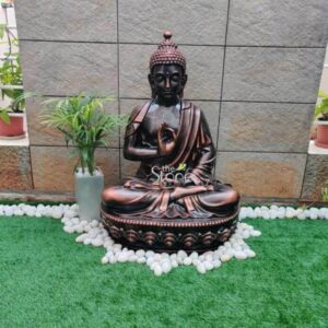 A serene 3.5-foot fibre Buddha statue is prominently displayed in what appears to be a well-maintained outdoor home garden setting. The statue with its rich bronze-like finish depicts Buddha seated in the classic lotus position or Padmasana signifying meditation and spiritual purity. His right hand is raised in the Vitarka Mudra a gesture symbolizing the discussion and transmission of Buddhist teachings while his left hand rests gently in his lap. The Buddha's face exudes tranquility with downcast eyes a gentle smile and elongated earlobes. The statue rests on a decorative circular base adorned with sculpted lotus petals further enhancing its spiritual aesthetic. Surrounding the base and extending onto the vibrant green artificial grass is a border of smooth white river stones creating a visually pleasing contrast. Behind the Buddha a tall slender potted plant with lush green leaves adds a touch of natural elegance. The background features a modern wall constructed from light grey rectangular stone tiles providing a contemporary backdrop for the traditional statue. This fibre Buddha statue with its ideal height and peaceful demeanor is perfectly suited for a home in Coimbatore as highlighted in the article. Its durable fibre construction makes it suitable for both indoor and outdoor placement while its artistic design and calming presence make it a focal point for meditation or simply as a decorative element. The harmonious arrangement of the statue the white stones and the potted plant creates an inviting and tranquil atmosphere embodying the essence of peace that a Buddha statue brings to a home.