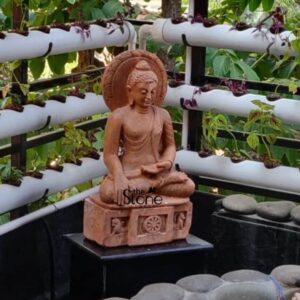 A serene Buddha statue rendered in a reddish-brown stone or terracotta-like material is beautifully situated in what appears to be a modern outdoor home environment possibly a balcony or terrace in Mumbai. The Buddha is depicted in a meditative seated posture likely Padmasana with his hands resting gently in his lap in a Dhyana Mudra symbolizing contemplation and spiritual awakening. His expression is one of profound peace and tranquility with eyes downcast and a subtle smile. The statue features a distinctive circular halo or aureole behind the Buddha's head intricately carved with radiating lines signifying his enlightened status. The Buddha sits upon a rectangular carved base that incorporates symbolic Buddhist imagery including the Dharma Chakra at its center flanked by smaller figures or motifs. This base rests on a sleek dark polished rectangular slab which in turn is positioned above a bed of smooth dark river stones creating a minimalist yet harmonious display. In the foreground more smooth grey stones are visible suggesting a contemplative rock garden element. The background of the image features a vertical hydroponic or vertical garden system with multiple white pipes each holding small green and purple-leafed plants adding a contemporary and refreshing touch to the setting. This Buddha statue with its contemplative posture and elegant design is perfectly suited for a home in Mumbai providing a calming focal point that blends traditional spirituality with modern living. The thoughtful arrangement of natural and artificial elements creates a peaceful sanctuary ideal for reflection and enhancing the home's ambiance.