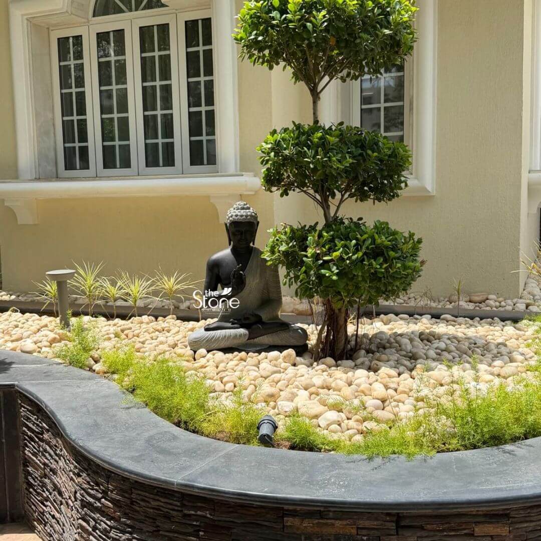 2.5ft Buddha Sculpture for Garden 6 A serene 2.5 Feet Blackstone Buddha Statue is elegantly placed in a meticulously designed modern garden likely in Gandhinagar Gujarat as suggested by its article context. This Buddha Stone Statue in Gandhinagar Gujarat features the Buddha seated in a meditative posture with hands resting in his lap palms upward. The statues dark blackstone material provides a striking contrast against the light-colored elements of the garden while its smooth polished surface reflects the tranquil ambiance. The Buddha's serene expression with closed eyes conveys profound peace and introspection making it a focal point for contemplation. The Buddha statue is situated amidst a bed of smooth light beige or off-white river stone which cover a substantial area of the garden. A meticulously sculpted bonsai-style tree with a tiered canopy of dense green leaves stands immediately to the right of the Buddha providing shade and a natural frame. Delicate green ground cover or small plants are interspersed among the pebbles and around the base of the tree adding subtle touches of greenery. In the background a light-colored building with white-framed windows indicates a residential or institutional setting. The garden design includes a curved dark grey stone retaining wall in the foreground built with stacked rough-hewn stones adding textural interest and defining the garden bed. Discreet outdoor lights are embedded in the ground suggesting that the area is beautifully illuminated at night further enhancing the statues presence. This detailed composition of the 2.5 Feet Blackstone Buddha Statue within a thoughtfully landscaped environment not only highlights its aesthetic appeal but also its role in creating a peaceful and spiritual sanctuary connecting the visual elements to an article about a Buddha Stone Statue in Gandhinagar Gujarat.