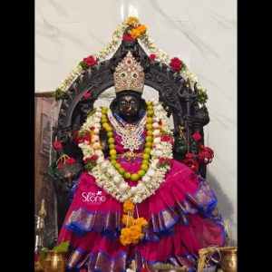 A stunning 43 inch Durga Devi Statue in Blackstone richly adorned and worshipped is presented in what appears to be a vibrant temple or shrine setting possibly in Nizamabad as suggested by its article context. This Durga Devi Statue in Nizamabad is intricately carved from dark blackstone depicting the powerful goddess Durga Devi seated regally. Her primary face is serene yet commanding with a powerful presence. The dark polished blackstone provides a striking contrast to her colorful adornments emphasizing her divine form. Durga Devi is draped in a beautiful flowing magenta-pink sari with intricate blue and gold borders which cascades gracefully around her seated form. She is magnificently adorned with multiple fresh flower garlands predominantly white and green beads intermingled with red roses and yellow marigolds symbolizing devotion and purity. A dazzling elaborate crown embellished with colorful jewels sits atop her head adding to her regal appearance. A detailed black stone archway or prabhavali decorated with traditional motifs and possibly a trident frames the goddess further enhancing her divine aura. At the base of the statue a collection of fresh flowers and golden brass offerings are placed reverently indicating active worship. The background features a white marble wall with subtle grey veining providing a clean yet elegant backdrop for the deity. The overall composition powerfully conveys Durga Devi's immense spiritual power beauty and protective nature. This detailed description effectively connects the visual elements of the 43 inch Durga Devi Statue in Blackstone with its spiritual significance and aesthetic qualities, making it an ideal representation for an article about a Durga Devi Statue in Nizamabad highlighting its role in inspiring devotion and strength.