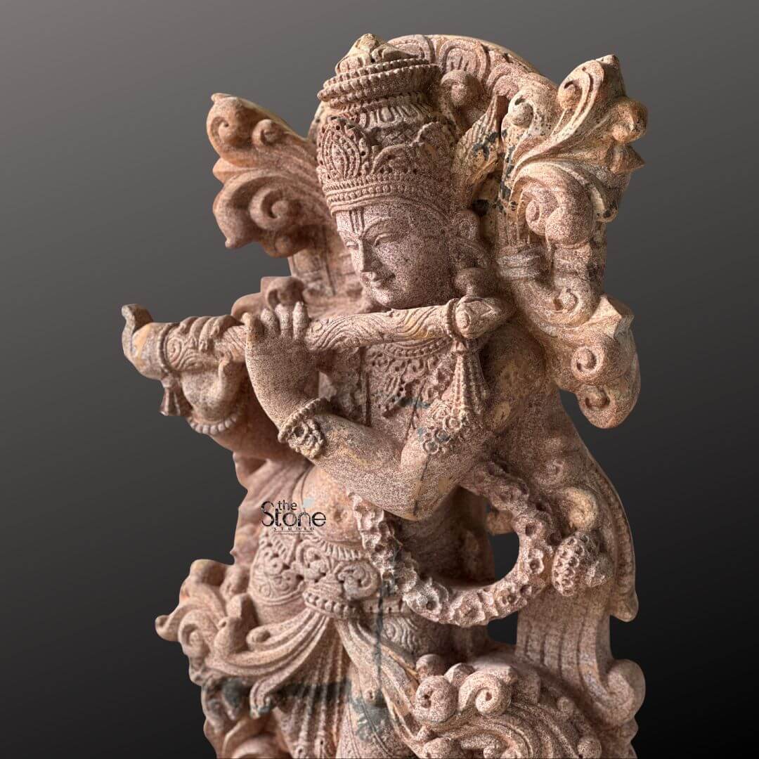 Standing Krishna Stone Statue with Flute 3.5ft 4 A close-up view captures the intricate details of a 3.5-foot Flute Krishna stone statue expertly carved from sandstone set against a deep contrasting background. Lord Krishna is depicted gracefully playing his bansuri with his head tilted slightly and a serene expression on his face evoking the enchanting melodies for which he is renowned. His hands are delicately positioned to hold the flute highlighting the focal point of this divine portrayal. The sandstone sculpture is a marvel of artistic detail particularly in its adornments. Krishna wears a highly elaborate crown intricately carved with patterns and possibly featuring a peacock feather a quintessential symbol associated with him. His body is bedecked with rich jewelry including a prominent necklace detailed armlets and a waist belt all rendered with precision in the stone. The textured surface of the sandstone emphasizes the fine lines and curves of the drapery around his waist and legs adding to the sculpture's tactile and visual appeal. An ornate swirling design forms a decorative frame behind his upper body possibly representing auspicious foliage or clouds. This detailed image of the 3.5-foot sandstone Flute Krishna statue is perfectly suited for an article focusing on such stone artistry. The warm earthy tones of the sandstone beautifully capture the divine charm of Krishna making the flute-playing pose come alive. The close-up perspective allows viewers to appreciate the masterful carving from the subtle expression on Krishna's face to the intricate patterns on his crown and jewelry underscoring the statue's spiritual and aesthetic significance.