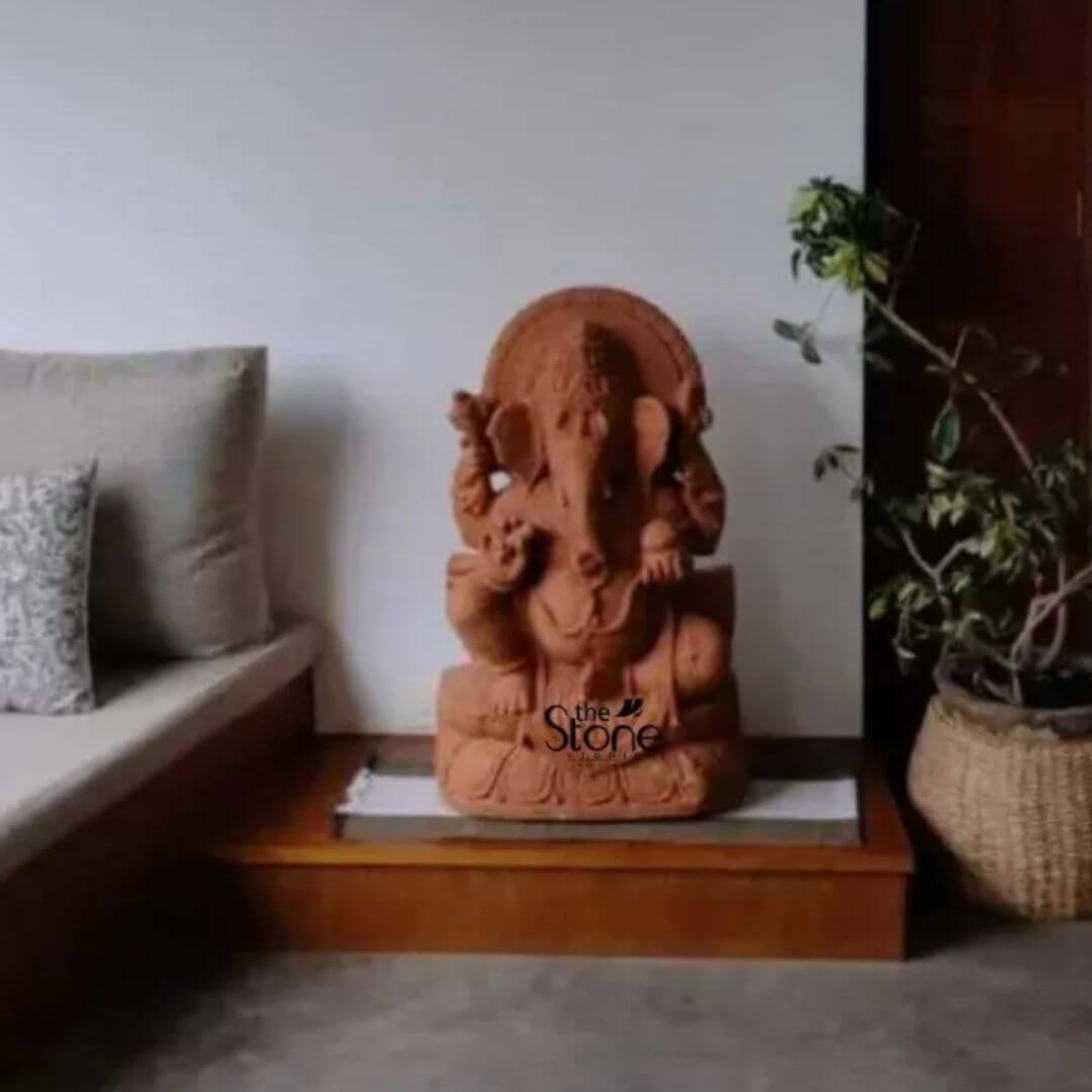 Ganapati Murti in Sand Stone 2.5' 4 A beautifully crafted 2.5-foot Ganesha Sandstone statue is prominently displayed on a dark wooden platform serving as a focal point in a well-appointed indoor setting likely in a home in Pondicherry. Lord Ganesha the elephant-headed god of new beginnings and remover of obstacles is depicted seated in a traditional posture exuding a sense of benevolence and wisdom. The sandstone material gives the statue a warm earthy reddish-brown hue. The Ganesha statue is rich with intricate details. Ganesha's iconic elephant head features large ears a prominent trunk and wise eyes. He is multi-armed holding his various attributes such as a modak a broken tusk and possibly an ankus and a paasha symbolizing his divine powers. He is adorned with sculpted jewelry and a crown all rendered with precision. A decorative arch or halo rises behind his head intricately carved with traditional motifs adding to the grandeur of the piece. The statue rests on a tiered base also carved with ornamental patterns including stylized lotus petals. This 2.5-foot sandstone Ganesha statue is an ideal subject for an article about Ganesha statues in Pondicherry highlighting its suitability for home altars or decorative displays. The warm tones of the sandstone create a welcoming and auspicious atmosphere. In the background hints of a cozy interior including a light-colored sofa with patterned cushions and a potted plant in a wicker basket suggest a comfortable living space. The entire arrangement contributes to a spiritual and aesthetically pleasing environment bringing blessings and positive energy into the home.