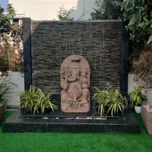 A impressive 5-foot Ganesha stone statue carved from sandstone prominently featured within a modern outdoor water fountain setup in Visakhapatnam as highlighted in the accompanying article. Lord Ganesha is depicted in a traditional seated posture against an arched backdrop his elephant head facing forward with a benevolent expression. The natural reddish-brown hue of the sandstone gives the statue an earthy and timeless aesthetic contrasting with the dark contemporary fountain elements. The Ganesha figure is richly detailed showcasing his four arms. His upper hands hold a goad and a noos while his lower right hand is raised in the Abhaya Mudra offering blessings. His lower left hand gently cradles a modak symbolizing abundance and joy. He is adorned with exquisitely carved jewelry including necklace armlets and a sacred thread across his chest all rendered in the sandstone. The arched backdrop behind him features traditional Hindu motifs and intricate carvings. The entire Ganesha statue is positioned at the base of a modern black stone waterfall feature where water gently cascades down a textured black wall behind it creating a serene and calming ambiance. The statue rests on a dark polished granite platform flanked by two potted spider plants adding a touch of greenery. The water collects in a narrow basin in front which is part of a larger dark granite rectangular base itself resting on artificial green turf. This setup represents a harmonious blend of traditional spiritual artistry with modern landscape design making it a significant focal point in Visakhapatnam.