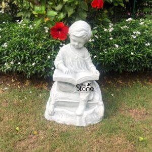 A charming 2-foot garden reading boy statue crafted from fiber and finished in a light stone-like texture is thoughtfully placed amidst a vibrant garden setting. The statue depicts a young boy sitting cross-legged engrossed in reading an open book exuding a sense of innocence and peaceful contemplation. The light color of the fiber statue stands out against the lush green grass in the foreground and the colorful foliage and flowers in the background making it a delightful focal point for any outdoor space. The boy is sculpted with soft gentle features his head slightly bowed as he concentrates on his book. His attire is simple with natural folds in the clothing adding to its realistic appearance. The book itself is open with pages and a binding clearly defined further enhancing the illusion of a child absorbed in a story. The texture of the fiber material mimics natural stone providing durability and a classic aesthetic suitable for outdoor display in places like Chandigarh where garden aesthetics are appreciated. This 2-foot Fiber Garden Reading Boy Statue is an enchanting addition to any garden evoking a sense of tranquility curiosity and the simple joys of childhood. Its durable fiber construction ensures longevity in outdoor conditions while its timeless design makes it a versatile piece that complements various garden styles. The statue not only adds a touch of artistic charm but also creates an inviting and serene atmosphere making it a beloved feature in a personal garden or public park.