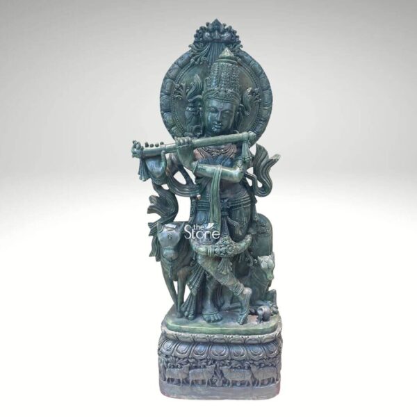 A magnificent Green Stone Krishna with Cow Statue stands prominently against a clean white background showcasing exquisite detail and artistry. Lord Krishna is depicted in his iconic Tribhanga pose, gracefully playing his flute his body angled with a serene expression. Beside him, a cow and a calf are intricately carved reflecting his identity as a divine cowherd an essential aspect of the Krishna with Cow theme. The greenstone material gives the entire sculpture a distinctive earthy and revered appearance. The greenstone Krishna statue is rich with traditional ornamentation. Krishna wears an elaborate crown likely adorned with a peacock feather a symbol strongly associated with him. His form is embellished with finely carved jewelry including necklaces armlets waistbands and anklets. The flowing drapery of his dhoti and sashes adds to the dynamic quality of the pose. Behind Krishna a circular aureole or halo frames his head often symbolizing divinity and a larger ornate arch rises above featuring intricate patterns. The cow and calf are rendered with gentle expressions symbolizing prosperity and devotion and they are surrounded by carved foliage and other natural elements. This Green Stone Krishna with Cow statue is an ideal visual for an article detailing such divine sculptures. The statue rests on a multi-tiered intricately carved base which features a band of smaller animal figures possibly more cows further enhancing the pastoral theme. The harmonious composition of Krishna the cow and the detailed base in greenstone creates a powerful and spiritually uplifting piece embodying devotion divine love and the sacred bond between Krishna and nature perfect for a temple or a home seeking a touch of spiritual grace.