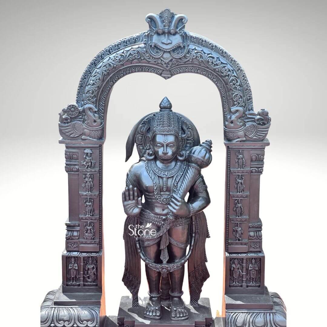 Standing Bajrangbali Statue 4ft 6 An impressive hand-carved Hanuman Murti sculpted from black stone and framed by an intricately decorative temple arch stands as a powerful and revered figure against a plain light background. This stunning black stone statue captures Lord Hanuman in a dynamic standing posture exuding an aura of immense strength devotion and courage perfectly complemented by its elaborate architectural setting. The dark polished surface of the black stone provides a dramatic contrast enhancing the intricate details of the sculpture and its ornate frame making it a profound focal point for worship. Lord Hanuman is depicted with his characteristic features including a majestic crown prominent ear ornaments and a strong muscular physique that symbolizes his extraordinary power. His right hand is raised in the Abhaya Mudra offering blessings and protection to devotees while his left hand firmly holds his Gada resting on his shoulder symbolizing his immense power and readiness to vanquish evil. A garland of beads adorns his neck and a decorative sash drapes across his body all carved with meticulous precision. He stands on a lotus pedestal which is integrated into a multi-tiered base adorned with smaller figures and decorative carvings all contributing to the grandeur of the piece. The intricate temple archway above him is equally detailed featuring divine figures floral motifs and a Kirtimukha at the apex flanked by mythical creatures. This hand-carved black stone Hanuman Murti with its decorative temple frame is a profound representation of Lord Hanuman the epitome of strength devotion and selfless service. The exceptional craftsmanship in black stone not only makes it a visually striking piece but also embodies deep cultural and religious significance offering a powerful focal point for devotion and prayer. The rich detailing of the deity and his surrounding archway reflects centuries of tradition making it a truly remarkable and revered artifact that brings blessings protection and a spiritual presence into any sacred space.