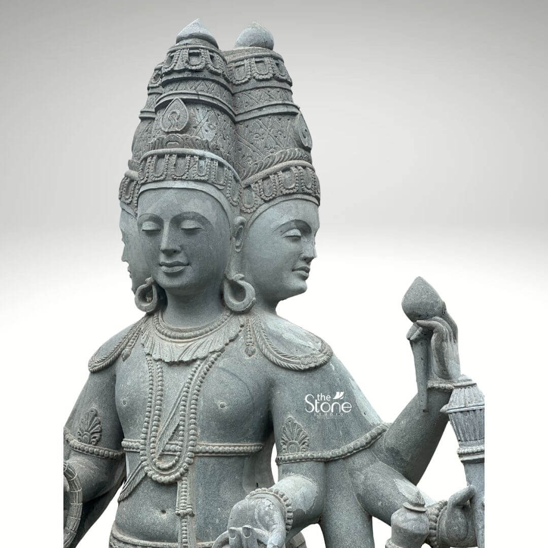 Brahma Stone Statue 7ft 8 A close-up high-detail image of a hand-carved 7-foot Greystone Lord Brahma Murti,focusing on the deity's upper body and characteristic four heads. The statue depicts Lord Brahma with serene and contemplative faces symbolizing his omnipresence and knowledge. The natural grey tones and subtle textures of the stone beautifully highlight the intricate details of the carving making it a profound and artistic representation of the creator god. Lord Brahma's four faces are meticulously sculpted each adorned with an elaborate tiered crown featuring intricate patterns. He is richly adorned with multiple necklaces, including beaded strands and larger decorative pieces along with armlets and a sacred thread all finely detailed. His multiple hands are shown holding his traditional attributes: one hand gently clasps a puskara while others hold a sruva or rosary. The fine craftsmanship is evident in the delicate facial features the texture of the crowns and the precise rendition of his ornaments showcasing the artisan's skill in transforming stone into a divine form. This hand-carved 7-foot Greystone Lord Brahma Murti is a profound representation of creation wisdom and cosmic knowledge. The exceptional artistry in natural greystone not only makes it a visually striking piece but also imbues it with deep cultural and religious significance. This exquisitely detailed Murti serves as a commanding focal point for devotion and praye radiating positive energy and ancient wisdom and is a testament to traditional stone carving ideal for a temple or a significant spiritual collection.