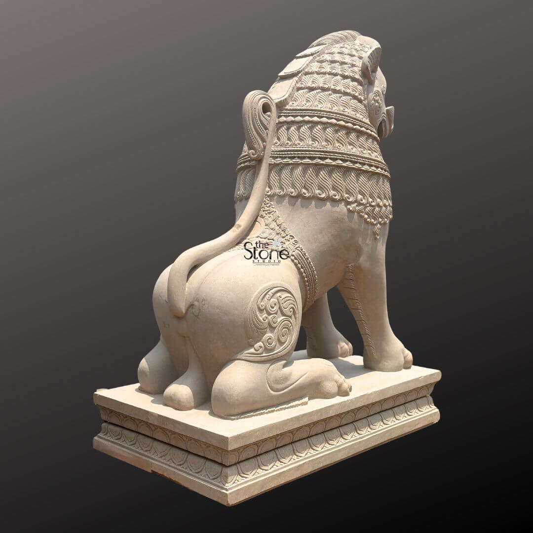 Traditional Stone Temple Lion Statue 7ft 2 A magnificent 7-foot hand-carved temple Simha statue sculpted from off-white sandstone is presented from a rear-right slightly elevated perspective against a dark plain background. This viewpoint emphasizes the lion's powerful physique and the intricate decorative carvings highlighting its role as a majestic guardian. The light natural hue of the sandstone allows the elaborate details to stand out vividly showcasing the exceptional craftsmanship involved in creating this traditional piece. The lion is depicted in a seated alert posture with its powerful hindquarters and robust legs prominently displayed. Its tail curls elegantly along its back terminating in an ornamental flourish a characteristic of traditional temple art. The mane is intricately carved with layers of textured patterns and decorative bands adorn its neck and shoulders. Swirling motifs and other traditional embellishments are visible on its flank and haunches adding depth and cultural richness to the sculpture. The entire sculpture rests upon a robust multi-tiered base also adorned with intricate patterns providing a solid foundation and enhancing its imposing presence. This 7-foot off-white sandstone Hand-Carved Temple Simha Statue is a profound representation of power protection and divine authority perfectly suited for the entrance of a temple or a significant architectural site. The exceptional craftsmanship in sandstone not only makes it a visually striking piece but also imbues it with deep cultural and religious significance. This exquisitely detailed statue serves as a commanding sentinel radiating strength and an ancient spiritual presence embodying centuries of artistic tradition and architectural grandeur.