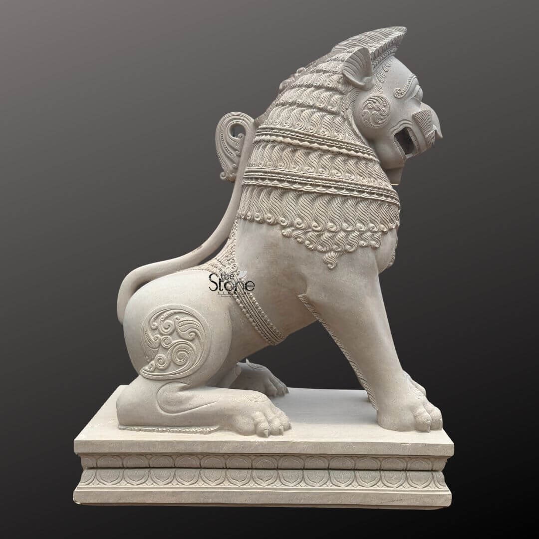Traditional Stone Temple Lion Statue 7ft 4 A magnificent 7-foot handcrafted stone lion statue sculpted from off-white sandstone is presented in a stately side profile against a dark plain background emphasizing its grandeur and intricate details. The statue depicts a powerful lion in a seated alert posture embodying strength guardianship and regal authority making it an impressive piece of art for grand entrances or gardens. The light natural hue of the sandstone allows the elaborate carvings to stand out vividly showcasing the exceptional artistry. The lion's features are highly stylized and ornamental typical of traditional stone carving. Its mane is intricately carved with layers of textured patterns resembling scales or braided hair and its powerful neck is adorned with decorative bands. The lion's face seen in profile features prominent eyes a strong snout and an open mouth conveying a sense of watchful guardianship and a roaring presence. Its muscular body is embellished with swirling motifs and decorative elements on its shoulders and hindquarters. The tail curls elegantly along its back terminating in an ornamental flourish. The entire sculpture rests upon a robust multi-tiered base also adorned with intricate patterns providing a solid foundation and enhancing its imposing presence. This 7-foot off-white sandstone Handcrafted Stone Lion Statue is a profound representation of powe protection and majesty suitable for a wide range of impressive architectural or landscape settings. The exceptional craftsmanship in sandstone not only makes it a visually striking piece but also imbues it with deep cultural and artistic significance. This exquisitely detailed statue serves as a commanding sentinel radiating strength and an ancient spiritual presence, embodying centuries of artistic tradition and skilled stone carving.