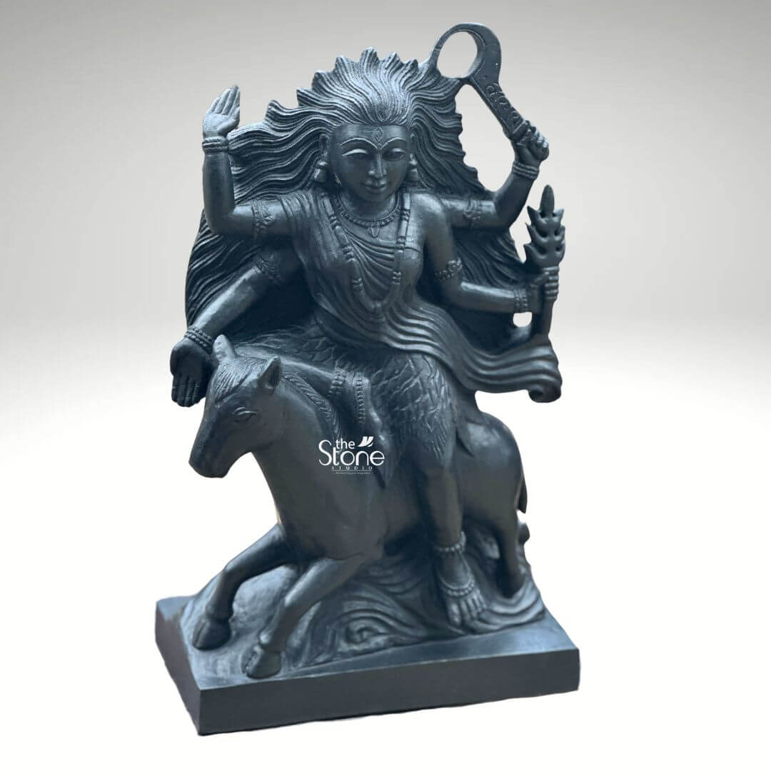 Black Stone Kaalratri Statue 15" 5 A powerful Kaalratri idol carved from black stone is dynamically posed and presented against a clean white background. This fierce manifestation of the Divine Mother one of the Navadurga is depicted riding a horse symbolizing her swiftness and power in overcoming malevolence. Her hair appears unbound and flows dramatically upwards and outwards emphasizing her formidable and untamed nature which aligns with her name meaning the night of blackness. The black stone Kaalratri idol is rich with symbolic and intricate details. The goddess is multi-armed with her hands wielding various weapons and ritual implements one hand prominently holds a saber or sword-like weapon aloft while another grasps a trident or a similar fiery object signifying her destructive power against evil. Her expression though intense conveys a sense of divine purpose and protective energy. She is adorned with sculpted jewelry including necklaces and bangles which stand out against her dark form. The horse she rides is captured mid-stride its legs seemingly galloping through stylized waves or clouds carved to enhance the sense of movement and raw power. The deep uniform black of the stone enhances the idol's dramatic and revered presence. This striking black stone Kaalratri idol is an excellent visual for an article detailing such powerful deities. The choice of black stone perfectly embodies her dark transformative aspect while the intricate carving of her wild hair weapons and dynamic pose on the horse conveys her relentless energy. The idol is a compelling representation of Kaalratri's role in vanquishing fear ignorance and negativity making it a significant piece for devotees and art enthusiasts seeking to understand her divine might and incorporate her protective presence.
