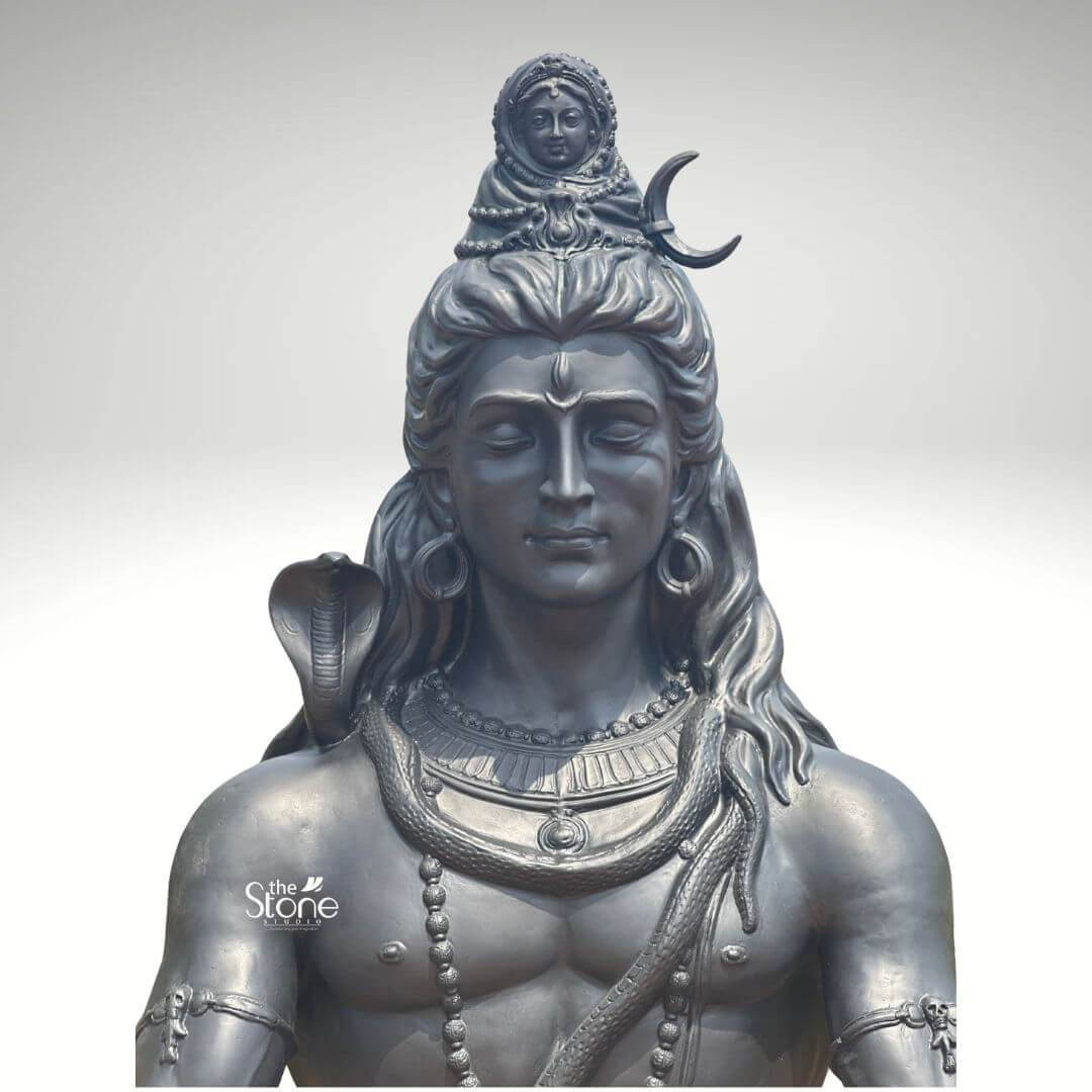 6 Feet Meditating Lord Shiva Statue 8 A close-up high-detail image of a large 6-foot Adiyogi Shiva Fibre Statue finished to resemble dark metal focuses on the deity's upper body and intricate iconography. This magnificent statue depicts Lord Shiva with his eyes gently closed in deep meditation radiating an aura of profound tranquility cosmic power and divine wisdom. As a large Adiyogi Shiva Fibre Statue it is an impressive and inspiring piece ideal for indoor decor showcasing meticulous craftsmanship and spiritual depth. Lord Shiva's face is sculpted with remarkable serenity featuring a prominent tilak on his forehead. His long flowing matted hair is elaborately piled high adorned with a crescent moon and a miniature figure of Goddess Ganga symbolizing the sacred river's descent. A lifelike cobra gracefully coils around his neck and multiple rudraksha bead malas drape across his chest and muscular torso along with other ornate necklaces and armbands. The dark metallic finish of the fiber material captures the subtle play of light and shadow highlighting every intricate detail of the carving from the texture of his hair to the smooth contours of his skin giving it a powerful contemplative presence. This large 6-foot Adiyogi Shiva Fibre Statue is a powerful representation of the ultimate reality the destroyer of evil and the source of all yoga and meditation perfectly suited for impactful indoor decor. The exceptional artistry in fiber allows for such intricate detailing and a commanding yet peaceful, presence making it a visually striking and spiritually significant artifact. This statue not only serves as a focal point for devotion and meditation but also as a majestic piece of art that radiates peace strength and cosmic energy enriching any space with its profound cultural and religious resonance truly embodying the spirit of Adiyogi.