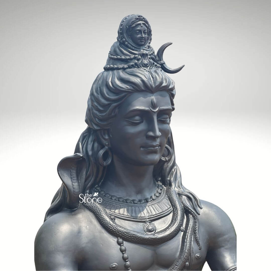 6 Feet Meditating Lord Shiva Statue 10 A close-up high-detail image of a large 6-foot Fibre Meditating Shiva Statue finished to resemble dark metal focuses on the deity's upper body and intricate iconography. This magnificent statue depicts Lord Shiva with his eyes gently closed in deep meditation radiating an aura of profound tranquility cosmic power and divine wisdom. As a large meditating Shiva statue it is an impressive and inspiring piece showcasing meticulous craftsmanship and spiritual depth. Lord Shiva's face is sculpted with remarkable serenity featuring a prominent tilak on his forehead. His long flowing matted hair is elaborately piled high adorned with a crescent moon and a miniature figure of Goddess Ganga symbolizing the sacred river's descent. A lifelike cobra gracefully coils around his neck and multiple rudraksha bead mala drape across his chest and muscular torso along with other ornate necklaces and armbands. The dark metallic finish of the fiber material captures the subtle play of light and shadow highlighting every intricate detail of the carving from the texture of his hair to the smooth contours of his skin giving it a powerful contemplative presence. This large 6-foot Fibre Meditating Shiva Statue is a powerful representation of the ultimate reality the destroyer of evil and the source of all yoga and meditation. The exceptional artistry in fiber allows for such intricate detailing and a commanding yet peaceful presence making it a visually striking and spiritually significant artifact. This statue not only serves as a focal point for devotion and meditation but also as a majestic piece of art that radiates peace strength and cosmic energy enriching any space with its profound cultural and religious resonance truly embodying the meditative aspect of Lord Shiva.