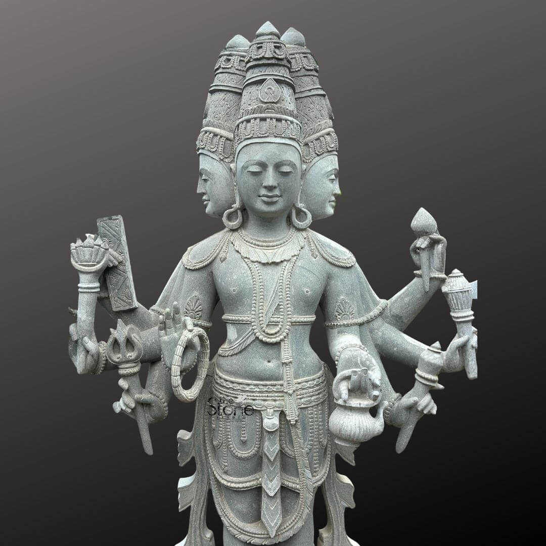 Brahma Stone Statue 7ft 9 A magnificent seven-foot tall statue of Lord Brahma meticulously carved from grey stone stands prominently against a dark featureless background. The deity is depicted with four serene faces each gazing in a different cardinal directio symbolizing his all-encompassing knowledge and presence. Each face is adorned with an intricate crown and their expressions exude a sense of calm wisdom. The statue's multi-armed form a hallmark of Hindu iconography is clearly visible with several hands holding various symbolic objects. The detailing on the grey stone Brahma statue is exquisite showcasing the artistry involved in its creation. Brahma is depicted holding a kamandalu a mala a pustaka and a sruk each representing different aspects of creation knowledge and ritual. The folds of his garments, the strands of his beaded necklaces and the bangles on his wrists are all carved with remarkable precision adding to the statue's lifelike quality. The grey tone of the stone enhances the statue's ancient and spiritual aura making it a compelling representation of the creator god. This impressive grey stone Brahma statue standing at an imposing seven feet perfectly encapsulates the grandeur and spiritual significance of Lord Brahma. Its detailed craftsmanship, from the multiple crowns to the symbolic objects held in each hand, makes it a powerful visual representation for an article discussing such divine artistry and religious symbolism. The soli, unyielding nature of the grey stone further emphasizes the eternal and foundational role of Brahma in Hindu cosmology.