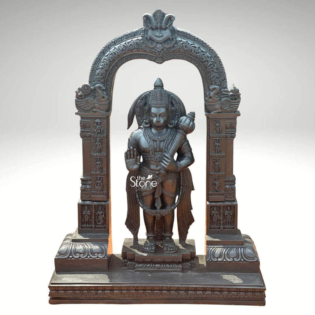 Standing Bajrangbali Statue 4ft 7 An impressive black stone Lord Hanuman statue framed by an intricately carved archway stands as a powerful and revered figure against a plain light background. This stunning black stone statue captures Lord Hanuman in a dynamic standing posture exuding an aura of immense strength devotion and courage. The dark polished surface of the stone provides a dramatic contrast enhancing the intricate details of the sculpture and its elaborate architectural frame making it a profound focal point for worship and admiration. Lord Hanuman is depicted with his characteristic features including a majestic crown prominent ear ornaments and a strong muscular physique that symbolizes his extraordinary power. His right hand is raised in the Abhaya Mudra offering blessings and protection to devotees while his left hand holds his Gada resting on his shoulder symbolizing his immense power and readiness to vanquish evil. A garland of beads adorns his neck and a decorative sash drapes across his body all carved with meticulous precision. He stands on a lotus pedestal which is integrated into a multi-tiered base adorned with smaller figures and decorative carvings all contributing to the grandeur of the piece. The intricate temple archway above him is equally detailed featuring divine figures floral motifs and a Kirtimukha at the apex flanked by mythical creatures like yalis or makaras. This black stone Lord Hanuman statue with its intricately carved arch is a profound representation of Lord Hanuman the epitome of strength devotion, and selfless service. The exceptional craftsmanship in black stone not only makes it a visually striking piece but also embodies deep cultural and religious significance offering a powerful focal point for devotion and prayer. The rich detailing of the deity and his surrounding archway reflects centuries of tradition making it a truly remarkable and revered artifact that brings blessings protection and a spiritual presence into any sacred space.