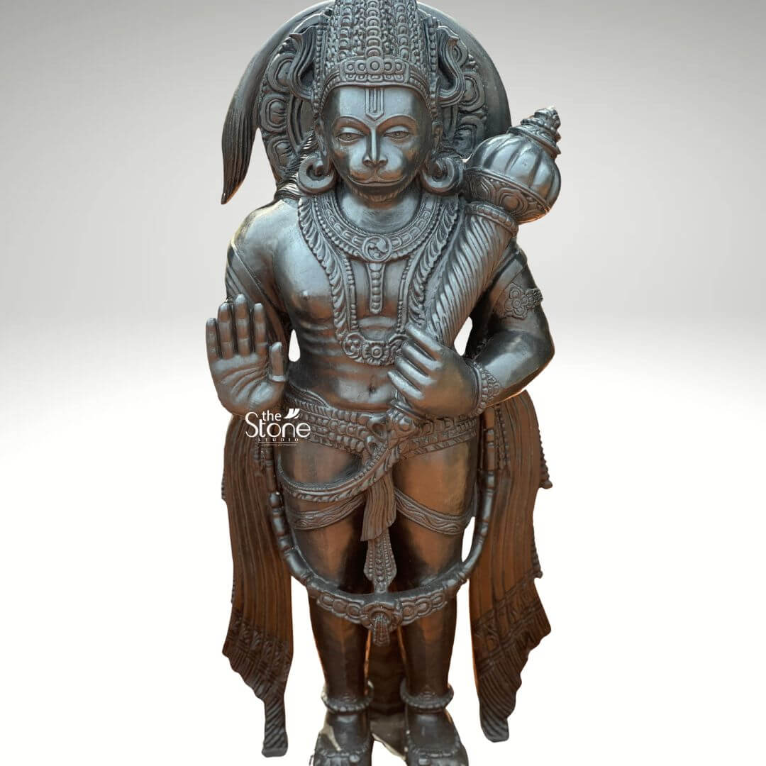 Standing Bajrangbali Statue 4ft 5 An impressive black stone Lord Hanuman idol depicted in the powerful Abhaya Mudra stands as a sole figure against a plain light background. This stunning black stone statue captures Lord Hanuman in a dynamic standing posture exuding an aura of immense strength unwavering devotion and courage. The dark polished surface of the stone provides a dramatic contrast enhancing the intricate details of the sculpture and making it a profound focal point for worship and reverence. Lord Hanuman is depicted with his characteristic features including a majestic crown prominent ear ornaments and a strong muscular physique that symbolizes his extraordinary power. His right hand is raised in the Abhaya Mudra a gesture offering blessings fearlessness and protection to devotees. His left hand firmly holds his Gada resting on his shoulder symbolizing his immense power and readiness to vanquish evil. A garland of beads adorns his neck and a decorative sash drapes across his body all carved with meticulous precision. He stands on a simple tiered pedestal conveying stability and divine presence. Behind his head, a subtle prabhavali or arch is suggested framing his divine form. This black stone Lord Hanuman idol in Abhaya Mudra is a profound representation of Lord Hanuman the epitome of strength devotion and selfless service. The exceptional craftsmanship in black stone not only makes it a visually striking piece but also embodies deep cultural and religious significance offering a powerful focal point for devotion and prayer. The rich detailing of the deity reflects centuries of tradition making it a truly remarkable and revered artifact that brings blessings protection and a spiritual presence into any sacred space.