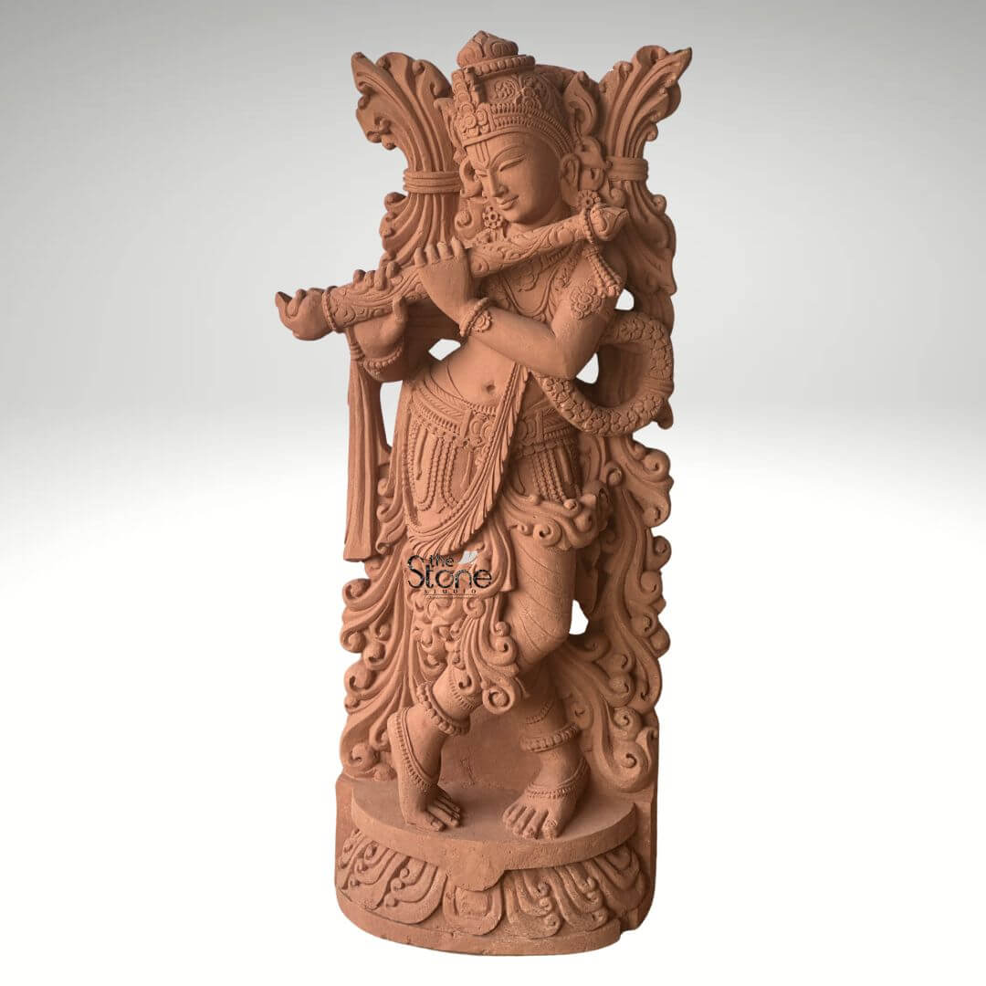 Lord Krishna Murti with Flute 3.5ft 4 A captivating 3.5-foot Lord Krishna standing statue meticulously carved from sandstone designed as a focal point for a temple as highlighted in the accompanying article. The entire statue rendered in the natural reddish-brown hue of sandstone exudes an earthy warm and timeless aesthetic. Lord Krishna is depicted standing in his iconic Tribhanga pose with one leg crossed gracefully in front of the other an expression of divine playfulness and charm. He holds a flute to his lips with both hands appearing to play an enchanting melody. The carving details on Krishna's attire including his dhoti sash and intricate jewelry such as necklaces armlets and ankle bracelets are exceptionally refined. He wears an elaborate crown and a garland or Vanamala drapes diagonally across his body. The flowing drapery of his garments is skillfully sculpted adding a sense of movement and grace to the figure. Behind Krishna an intricately carved design possibly representing foliage or an artistic prabhavali further enhances the visual richness of the murti providing a dignified frame. The sculpture stands on a circular ornate pedestal also crafted from sandstone featuring traditional Indian architectural motifs and decorative patterns adding to its grandeur and stability within a temple setting. The texture of the sandstone is visible throughout providing depth and authenticity to the artwork. This 3.5-foot sandstone Lord Krishna standing statue is a powerful representation of devotion and artistic mastery embodying the spiritual and aesthetic value discussed in the article making it an ideal centerpiece for spiritual veneration in a temple.