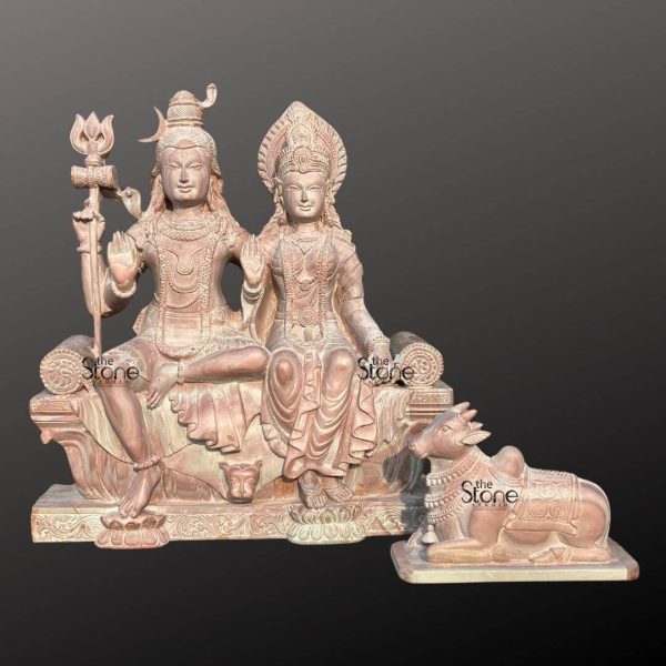 A beautiful 18-inch Lord Shiva Parvati statue crafted from pink stone accompanied by an 8-inch Nandi statue as highlighted in an article about such sculptures. The entire set rendered in a soft warm pinkish-brown hue of natural stone exudes a gentle and serene aesthetic. Lord Shiva and Goddess Parvati are depicted seated side-by-side on an ornate throne. Lord Shiva on the left has his traditional matted hair adorned with a crescent moon and a small cobra. He holds a trishul in his left hand while his right hand is raised in a blessing gesture. He wears minimal attire and intricate jewelry all carved from the stone. Goddess Parvati sits to Shiva's right adorned with an elaborate crown and traditional jewelry including necklaces and earrings. Her right hand is also raised in a blessing gesture while her left hand rests gracefully in her lap. Both deities have serene expressions conveying peace and divine harmony. The throne they share is intricately carved with traditional Indian motifs including a lion's face at the front symbolizing strength and royalty. The details on their garments flowing gently are also meticulously sculpted. Separately positioned to the right of the main statue is an 8-inch Nandi statue Lord Shiva's sacred bull also crafted from the same pink stone. Nandi is depicted in a reclining posture facing the divine couple adorned with carved blankets and jewelry symbolizing unwavering devotion. The smooth contours and fine details throughout all three figures emphasize a deep sense of tranquility and spiritual harmony. This Shiva Parvati and Nandi ensemble in its unique pink stone is a powerful representation of devotion and artistic mastery ideal for home altars or as a cherished decorative piece as discussed in the accompanying article.