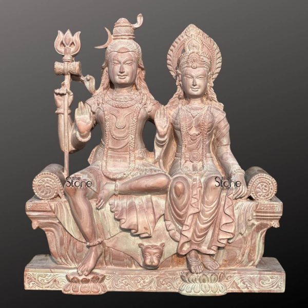 A majestic 18-inch Lord Shiva with Maa Parvati stone statue meticulously carved from pink stone presented against a dark background as highlighted in an article about such sculptures. The entire piece rendered in a soft warm pinkish-brown hue of natural stone exudes a gentle and serene aesthetic. Lord Shiva and Goddess Parvati are depicted seated side-by-side on an ornate throne. Lord Shiva on the left is depicted with his traditional matted hair adorned with a crescent moon and a small cobra. He holds a trishul in his left hand while his right hand is raised in the Abhaya Mudra. He wears minimal attire and intricate jewelry all carved from the stone. Goddess Parvati sits gracefully to Shiva's right adorned with an elaborate crown and traditional jewelry including necklaces and earrings. Her right hand is also raised in a blessing gesture while her left hand rests elegantly in her lap. Both deities have serene expressions conveying peace and divine harmony. The throne they share is intricately carved with traditional Indian motifs including a lion's face at the front symbolizing strength and royalty. The flowing drapery of their garments is also finely sculpted adding a sense of detail and elegance. The statue rests on a robust and ornate base also crafted from pink stone featuring lotus petal designs and other decorative patterns. The smooth contours and fine details throughout both figures emphasize a deep sense of tranquility and spiritual harmony. This Shiva Parvati stone sculpture in its unique pink stone is a powerful representation of devotion and artistic mastery making it an ideal piece for spiritual veneration and artistic appreciation in a home or temple as discussed in the accompanying article.