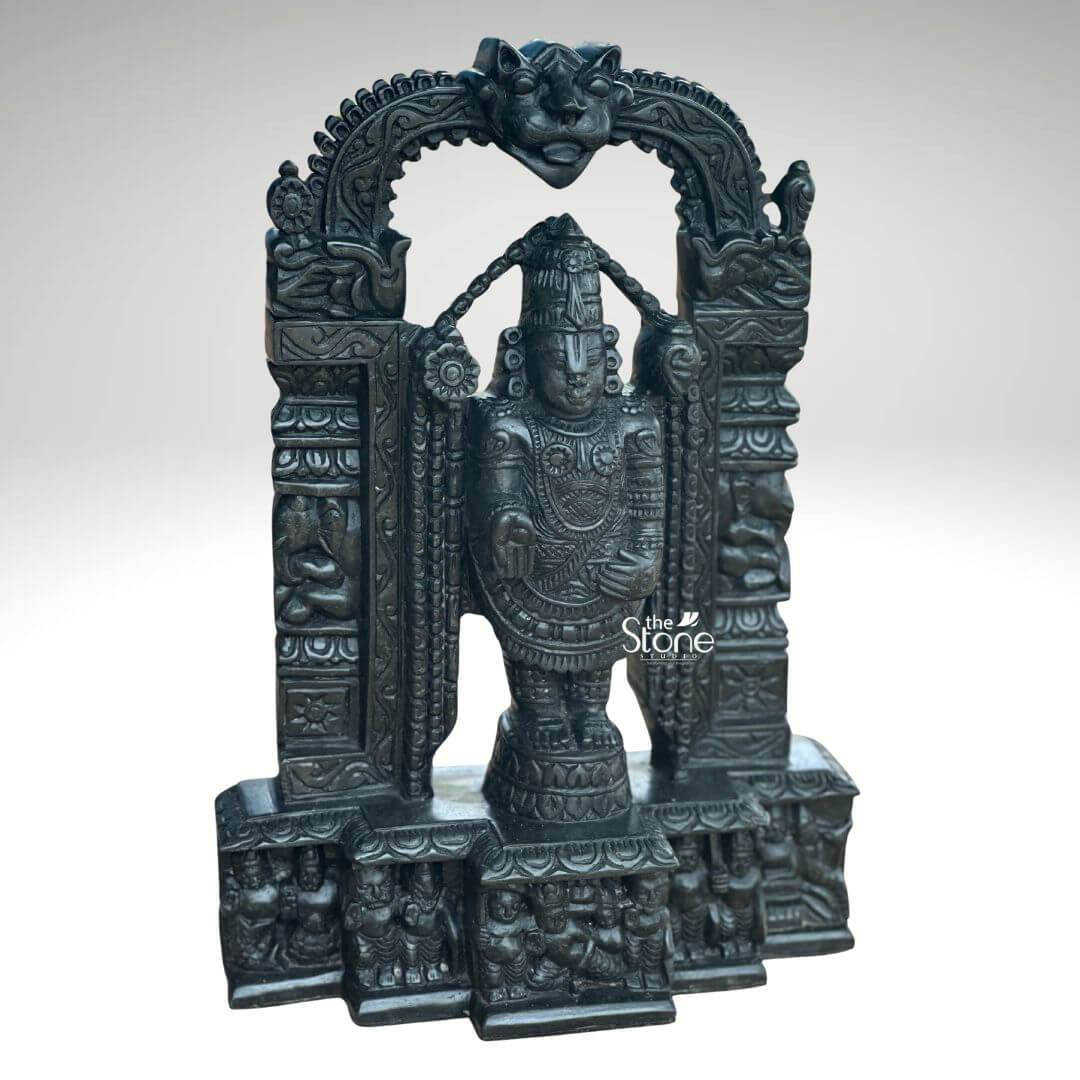 Tirupati Balaji Statue in Black Stone 18" 5 An exquisitely carved 18-inch Lord Srinivasa stone statue complete with an integrated temple arch is showcased against a plain light background. This stunning black stone Balaji statue captures Lord Srinivasa in his iconic standing posture emanating an aura of divine grace and profound power making it a perfect piece for a home temple or a significant spiritual collection. The deep polished surface of the black stone provides a dramatic contrast enhancing the intricate details of the sculpture and its ornate architectural frame. Lord Srinivasa is depicted with his characteristic features including the prominent Urdhva Pundra on his forehead a majestic crown and various traditional ornaments such as multiple necklaces armlets and a sacred thread all finely detailed. His right hand is raised in the Abhaya Mudra offering blessings and reassurance to devotees while his left hand rests on his thigh symbolizing protection and prosperity. He stands on a lotus pedestal which is integrated into a multi-tiered base adorned with smaller figures and decorative carvings including devotees and mythological scenes all contributing to the grandeur of the piece. The intricate temple archway above him is equally detailed featuring divine figures floral motifs and a Kirtimukha at the apex. This 18-inch black stone Lord Srinivasa statue with its integrated temple arch is a profound representation of Lord Venkateswara the preserver of the universe. The exceptional craftsmanship in black stone not only makes it a visually striking piece but also embodies deep cultural and religious significance offering a powerful focal point for devotion and prayer. The rich detailing of the deity and his surrounding archway reflects centuries of tradition making it a truly remarkable and revered artifact that brings blessings and a spiritual presence into any sacred space.