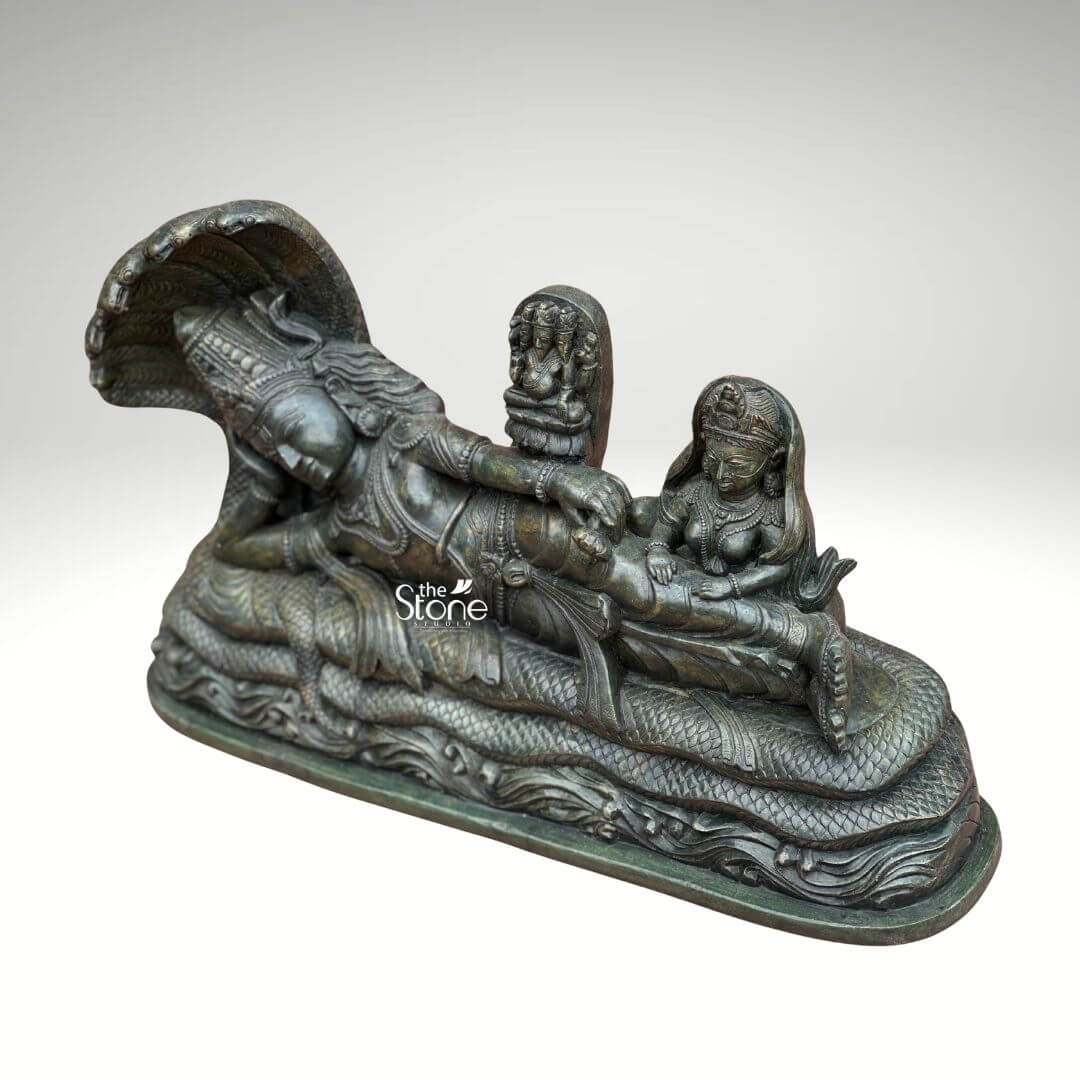 Ananta Shayana Vishnu Statue 20" 8 A majestic green stone statue captures the iconic depiction of Lord Vishnu sleeping on the cosmic serpent Sheshnag a profound representation of cosmic cycles and preservation in Hindu mythology. The sculpture presented from a slightly elevated oblique angle highlights the intricate details and the serene narrative of Vishnu's Ananta Shayana posture. The deep earthy green hue of the stone possibly chlorite schist or a similar dark fine-grained material enhances the ancient and sacred aesthetic lending a sense of timelessness to the detailed craftsmanship. Lord Vishnu is carved in a state of serene cosmic slumber his head gently supported by his right arm with an expression of ultimate tranquility. He is lavishly adorned with traditional Hindu ornaments including an elaborate crown multiple necklaces intricate armlets and a gracefully draped dhoti all carved with meticulous precision. The multi-headed Sheshnag coils beneath him its numerous hoods fanning out to form a grand protective canopy over Vishnu's head with its scales and undulating body intricately rendered. From Vishnu's navel a delicate lotus emerges bearing a miniature figure of Brahma the creator god symbolizing the universe's cyclical creation. To Vishnu's right seated attentively is Goddess Lakshmi his divine consort her form equally adorned and sculpted with reverence massaging his feet. This green stone statue of Lord Vishnu sleeping on Sheshnag is a powerful and sacred representation of preservation cosmic order and the cyclical nature of time. The exceptional detail and the choice of green stone often associated with spiritual balance and renewal enhance its profound symbolism. This exquisitely crafted statue is an impressive and deeply meaningful artifact ideal for a grand temple a prominent home altar or a spiritual garden inviting contemplation and reverence for the divine cosmic dance and enriching any space with its deep cultural and religious resonance.