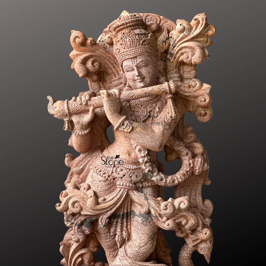 Standing Krishna Stone Statue with Flute 3.5ft 7 A close-up view highlights the intricate details of a 3.5-foot Murali Krishna stone statue expertly carved from sandstone set against a deep contrasting background. Lord Krishna is depicted gracefully playing his bansuri also known as Murali with his head tilted slightly and a serene contemplative expression on his face. His hands are delicately positioned to hold the flute making it the central focus of this divine portrayal emphasizing his identity as Muralidhara the flute-bearer. The sandstone sculpture is a marvel of artistic detail particularly in its rich ornamentation. Krishna wears a highly elaborate crown intricately carved with patterns and possibly featuring a peacock feather a quintessential symbol associated with him. His body is bedecked with rich jewelry including a prominent necklace detailed armlets and a decorative waist belt all rendered with precision in the stone. The textured surface of the sandstone emphasizes the fine lines and curves of the drapery around his waist and legs adding to the sculpture's tactile and visual appeal. An ornate swirling design forms a decorative frame behind his upper body possibly representing auspicious foliage or clouds. This detailed image of the 3.5-foot sandstone Murali Krishna statue is perfectly suited for an article focusing on such specialized stone artistry. The warm earthy tones of the sandstone beautifully capture the divine charm of Krishna making the flute-playing pose come alive. The close-up perspective allows viewers to appreciate the masterful carving from the subtle expression on Krishna's face to the intricate patterns on his crown and jewelry underscoring the statue's spiritual and aesthetic significance as a representation of Murali Krishna.