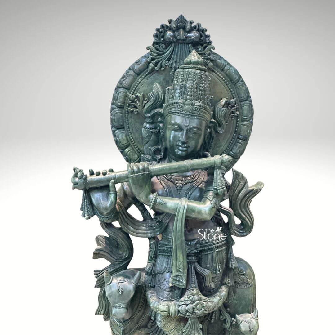 Green Stone Krishna with Cow Statue 4ft 6 A majestic Murli Krishna Green Stone Idol carved from a single piece of green stone stands prominently against a clean white background. The statue depicts Lord Krishna in his iconic Tribhanga pose with a gentle sway to his body. He is adorned with an elaborate crown featuring intricate carvings and delicate jewelry graces his neck arms and ankles. His serene face gazes forward eyes downcast in a posture of divine meditation as he holds a meticulously detailed flute to his lips poised to play. The craftsmanship is evident in the flowing drapery of his garments which are intricately sculpted to show texture and movement. A halo-like circular aureole with ornate patterns frames his head further emphasizing his divine presence. At his feet a small docile cow or calf is depicted a common motif associated with Krishna symbolizing his role as a protector and caretaker. The green stone itself exhibits variations in shade and subtle veining adding depth and character to the idol and highlighting the natural beauty of the material. This detailed Greenstone statue of Krishna playing the flute perfectly illustrates the artistry and spiritual significance discussed in the article about Murli Krishna Green Stone Idols. The idol's rich detailing, from the divine ornaments to the serene expression and symbolic cow makes it a captivating representation of the deity suitable for worship or as a decorative piece in a home or temple.