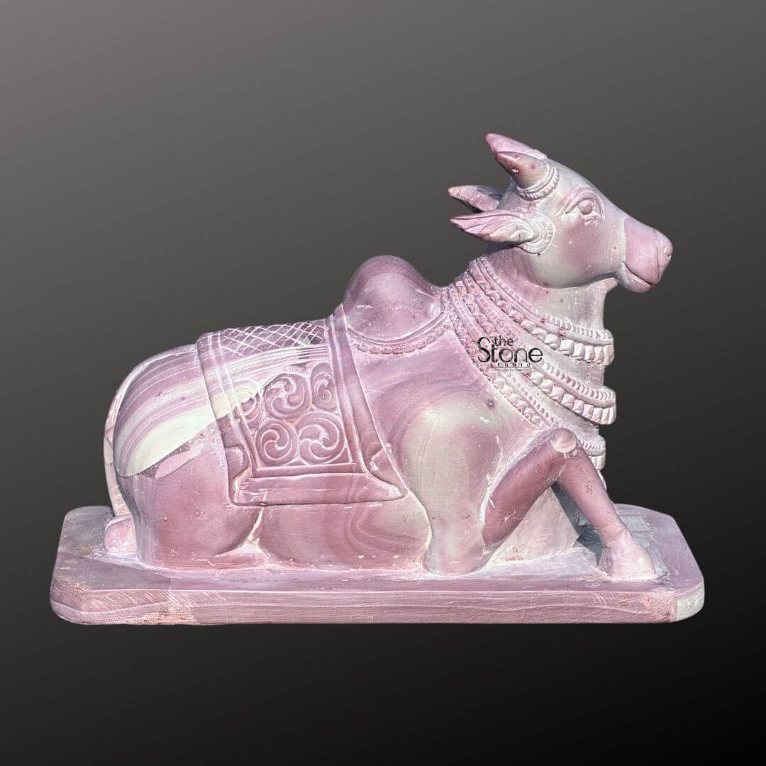 Stone Nandi Statue for Home Temple 8" 2 An exquisite 8-inch Nandi Bull statue meticulously carved from pink stone presented against a dark background as highlighted in an article about such sculptures. The entire sculpture rendered in a soft warm pinkish-brown hue of natural stone features beautiful natural veining and color variations giving it a unique and serene aesthetic. Nandi Lord Shiva's sacred bull is depicted in a traditional reclining posture symbolizing unwavering devotion patience and strength. The statue showcases remarkable detail in its carving. Nandi is adorned with a decorative saddle or blanket intricately carved with traditional Indian motifs and numerous strands of jewelry around his neck and head. His horns are neatly sculpted and his ears are gently laid back conveying a sense of calm and attentiveness. The eyes are subtly defined adding to the serene expression. The natural texture and subtle color variations of the pink stone are prominent throughout adding authenticity and depth to the artwork. The Nandi statue rests on a simple rectangular base also carved from the same pink stone ensuring stability and a cohesive look. The smooth contours and fine details emphasize a deep sense of tranquility and spiritual harmony. This 8-inch pink stone Nandi Bull statue is a powerful representation of devotion and artistic mastery perfectly suited for spiritual veneration in a home temple or as a cherished decorative piece embodying the revered companion of Lord Shiva as discussed in the accompanying article.
