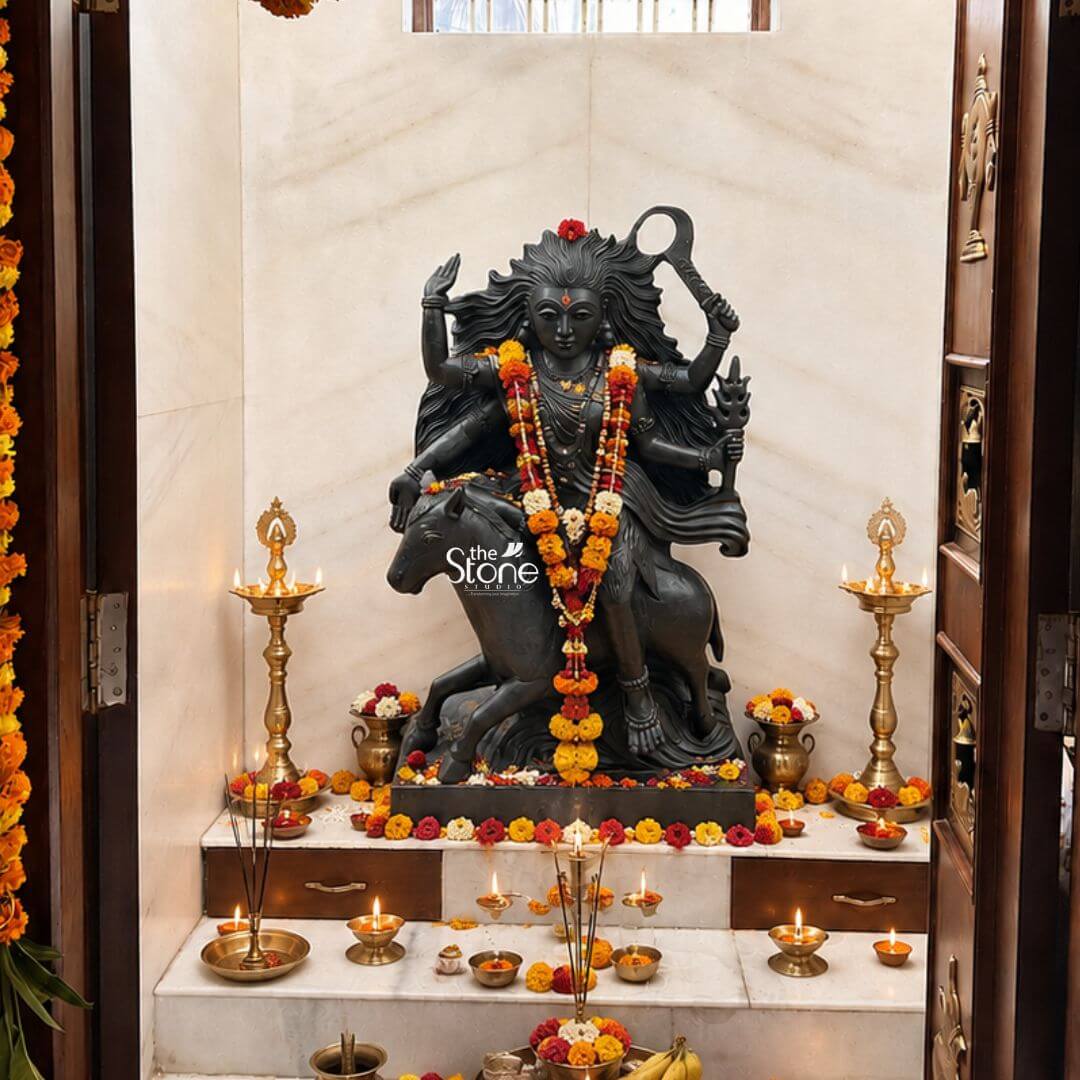 Black Stone Kaalratri Statue 15" 3 A majestically adorned Navadurga Kaalratri statue carved from black stone is the vibrant centerpiece of a beautifully decorated home temple likely during a festive occasion. The fierce manifestation of the Divine Mother is depicted riding a horse symbolizing her swift action and power against evil. Her wild flowing hair intense expression and multi-armed form are clearly visible enhanced by the devotional setting. The black stone statue of Kaalratri is lavishly garlanded with fresh orange and yellow marigold flowers draped around her neck and across the horse signifying deep reverence. Her multiple hands wield various symbolic weapons including a prominent sword or saber and a trident-like object. The statue is surrounded by a multitude of traditional Indian offerings numerous small brass diyas emit a warm glow creating a sacred ambiance. Bowls filled with fresh flowers kumkum and incense sticks are arranged meticulously on the steps leading up to the deity signifying prayers and devotion. Two tall, ornate brass lamps stand symmetrically on either side of the statue further illuminating the space. This striking black stone Navadurga Kaalratri statue beautifully decorated and situated within a home temple is an excellent visual for an article detailing such divine forms. The rich contrast of the dark stone against the vibrant marigold garlands and the warm glow of the diyas creates a powerful and spiritual atmosphere. The entire arrangement from the offerings to the lighting emphasizes the reverence and devotion surrounding Kaalratri making it a compelling representation of her protective energy and importance in Hindu worship.