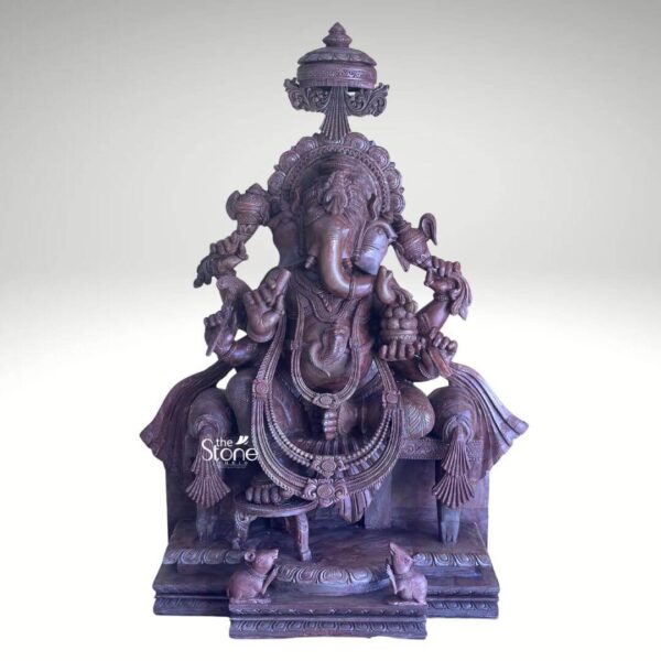A majestic 33-inch pink stone Ganesh idol is presented in a stately front-facing view against a plain light background emphasizing its grandeur and intricate craftsmanship. The statue depicts Lord Ganesha seated regally on a throne adorned with a magnificent crown and holding his traditional attributes exuding an aura of wisdom prosperit and divine blessings. The natural pink hues and subtle variations in the stone lend a warm inviting and auspicious quality to this impressive Ganesha statue. Lord Ganesha is depicted with his characteristic elephant head large ears and a gently curved trunk. He is adorned with an elaborate crown necklaces armlets and a sacred thread all meticulously carved with fine details. In his multiple hands he holds various implements such as an ankush a pasha and a modak his favorite treat. The throne he sits upon is also richly decorated with ornate carvings on its legs and backrest and beneath him his vehicle a small mouse is depicted reverently. A grand intricately carved archway or prabhavali frames the deity adding to his divine presence. This 33-inch pink stone Ganesh idol is a profound representation of wisdom prosperity and obstacle removal making it a powerful and auspicious addition to any home temple or significant spiritual space. The exceptional craftsmanship in natural pink stone not only makes it a visually striking piece but also imbues it with deep cultural and religious significance. This exquisitely detailed statue serves as a commanding focal point for devotion and prayer radiating positive energy and blessings to all who venerate it.
