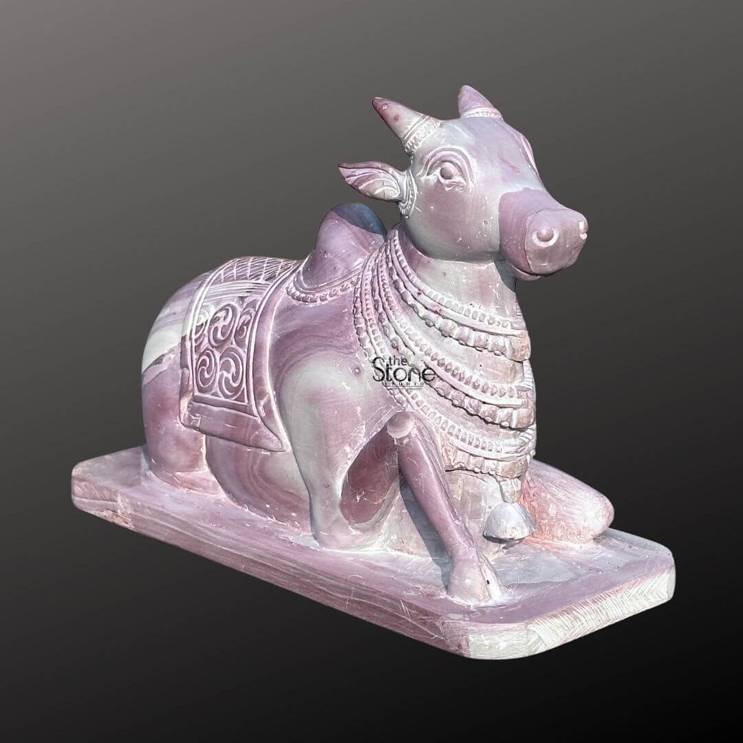 Stone Nandi Statue for Home Temple 8" 4 An exquisite 8-inch Pink Stone Nandi Idol meticulously carved from pink stone presented against a dark background making it an ideal piece for a Shiv temple as highlighted in the accompanying article. The entire sculpture rendered in a soft warm pinkish-brown hue of natural stone features beautiful natural veining and color variations giving it a unique and serene aesthetic. Nandi Lord Shiva's sacred bull is depicted in a traditional reclining posture symbolizing unwavering devotion patience and strength. The statue showcases remarkable detail in its carving. Nandi is adorned with a decorative saddle or blanket intricately carved with traditional Indian motifs and numerous strands of jewelry around his neck and head. A small bell is also visible on his chest. His horns are neatly sculpted and his ears are gently laid back conveying a sense of calm and attentiveness. The eyes are subtly defined adding to the serene expression. The natural texture and subtle color variations of the pink stone are prominent throughout adding authenticity and depth to the artwork. The Nandi idol rests on a simple rectangular base also carved from the same pink stone ensuring stability and a cohesive look. The smooth contours and fine details emphasize a deep sense of tranquility and spiritual harmony. This 8-inch pink stone Nandi idol is a powerful representation of devotion and artistic mastery perfectly suited for spiritual veneration in a Shiv temple embodying the revered companion and gatekeeper of Lord Shiva as discussed in the accompanying article.
