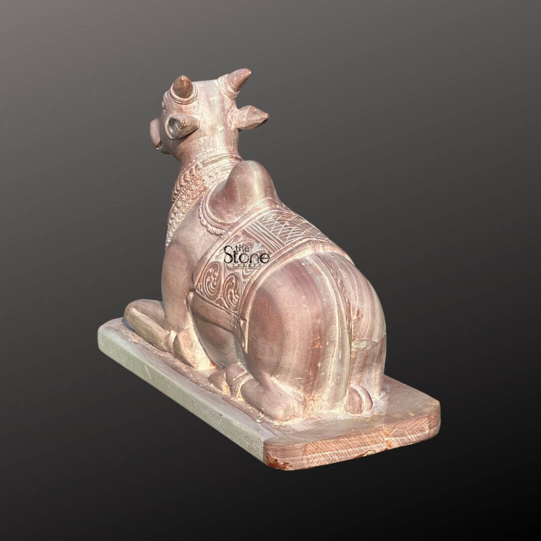 Stone Nandi Statue for Home Temple 8" 5 A beautifully carved 8-inch pink stone Nandi statue is depicted from a slightly elevated rear-left perspective showcasing its intricate details and connection to traditional Hindu worship. The statue features the sacred bull Nandi in a seated alert posture its head gently turned to the left and its powerful body sculpted with grace and reverence. The natural pink hue of the stone is subtly highlighted emphasizing the smooth polished surfaces and the skilled craftsmanship that brings the divine animal to life making it a fitting centerpiece for a puja room. The Nandi's back is adorned with an elaborately carved blanket featuring geometric patterns and swirling organic motifs that add depth and texture to the sculpture. Its humped back a characteristic of sacred Indian bulls is prominent and the musculature of its hindquarters is delicately rendered suggesting strength and devotion. The statue rests upon a simple rectangular base providing stability and a sense of grounding for the divine figure while its small size makes it ideal for personal altars and home temples. This exquisite pink stone Nandi statue a symbol of devotion strength, and faithfulness in Hindu mythology is perfectly suited for a puja room inviting a sense of tranquility and spiritual connection. The delicate pink stone radiates a serene energy complementing the sacred atmosphere of worship and meditation. The detailed carving and thoughtful design of this 8-inch Nandi make it not just a decorative item but a meaningful artifact for those seeking to enhance their spiritual practice and bring blessings into their home.