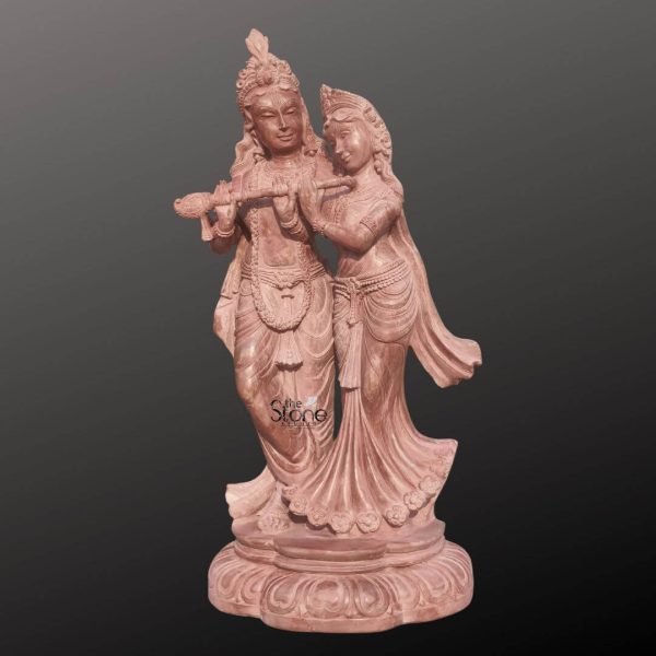 A captivating 20 inch Radha Krishna Stone Sculpture meticulously carved from beautiful pinkstone stands gracefully against a plain dark background highlighting its intricate details and loving depiction. This Radha Krishna Stone Sculpture portrays Lord Krishna and Radha his beloved consort in an enchanting embrace symbolizing divine love and harmony. Both figures are rendered with exquisite detail their gentle expressions conveying deep affection. The warm soft hue of the pinkstone gives the statue an ethereal and romantic quality making it a truly enchanting piece. Lord Krishna is depicted on the left in his iconic Tribhanga pose playing the flute. He wears a majestic crown with a peacock feather elaborate jewelry and flowing garments all finely sculpted. Radha on the right leans tenderly into Krishna her hand delicately touching his flute her gaze soft and adoring. She is adorned with a beautiful crown intricate jewelry and graceful drapery which flows elegantly around her form. The meticulous carving captures the delicate features of their faces their intertwined postures and the intricate details of their ornaments and clothing. The divine couple stands on a beautifully carved lotus-petal shaped base also made of pinkstone providing a stable and aesthetically pleasing foundation. The entire composition emanates a sense of divine romance devotion and artistic elegance. This detailed depiction of the 20 inch Radha Krishna statue in Pinkstone beautifully captures its spiritual essence and aesthetic appeal making it an ideal representation for an article about a Radha Krishna Stone Sculpture emphasizing its role in inspiring love and devotion.