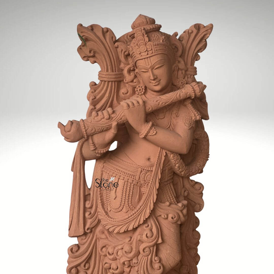 Lord Krishna Murti with Flute 3.5ft 5 A beautiful 3.5-foot sandstone Krishna statue explicitly designed for outdoor display as highlighted in the accompanying article. The entire sculpture rendered in the natural reddish-brown hue of sandstone exudes an earthy warm and timeless aesthetic that harmonizes beautifully with natural outdoor environments. Lord Krishna is depicted standing in his iconic Tribhanga pose with one leg crossed gracefully in front of the other exuding divine playfulness and charm. He holds a flute to his lips with both hands appearing to play an enchanting melody. The carving details on Krishna's attire including his dhoti sash and intricate jewelry such as necklaces armlets and ankle bracelets are exceptionally refined. He wears an elaborate crown and a garland or Vanamala drapes diagonally across his body. The flowing drapery of his garments is skillfully sculpted adding a sense of movement and grace to the figure. Behind Krishna an intricately carved design possibly representing foliage or an artistic prabhavali further enhances the visual richness of the murti. The sculpture stands on a circular ornate pedestal also crafted from sandstone featuring traditional Indian architectural motifs and decorative patterns adding to its grandeur and stability in an outdoor setting. The robust nature of sandstone makes this 3.5-foot Krishna statue a durable and powerful representation of devotion and artistic mastery perfectly suited to withstand outdoor elements while enhancing the beauty and tranquility of any garden, courtyard or open-air spiritual space as discussed in the article.