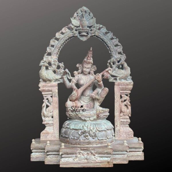 A stunning 20-inch Saraswati Stone Statue with Veena meticulously carved from pink stone is presented against a deep dark background. The goddess of knowledge music and art Saraswati is depicted seated gracefully in Lalitasana one leg folded the other dangling on a lotus base playing her sacred musical instrument the veena. Her serene expression and graceful posture exude divine wisdom and artistic harmony. The pink stone statue features an elaborate arch that frames the deity adding to its grandeur. This arch is intricately carved with celestial motifs including stylized birds likely peacocks or hamsas symbols of Saraswati at its corners and delicate scrolling patterns. Saraswati is multi-armed with two main hands positioned to play the veena while other hands hold symbolic items such as a mala and a pustaka representing her dominion over knowledge and meditation. Her intricate crown jewelry and flowing garments are all carved with exceptional detail highlighting the artisan's skill. The warm subtle pink hue of the stone adds a gentle and inviting quality to the piece. This 20-inch pink stone Saraswati statue with Veena is an ideal subject for an article discussing such divine sculptures. The detailed craftsmanship from the delicate strings of the veena to the intricate patterns on the arch and her adornments showcases a high level of artistry. The statue beautifully embodies the essence of Saraswati as the patron of arts and learning making it a perfect addition to a home educational institution or temple inspiring creativity wisdom and spiritual devotion.