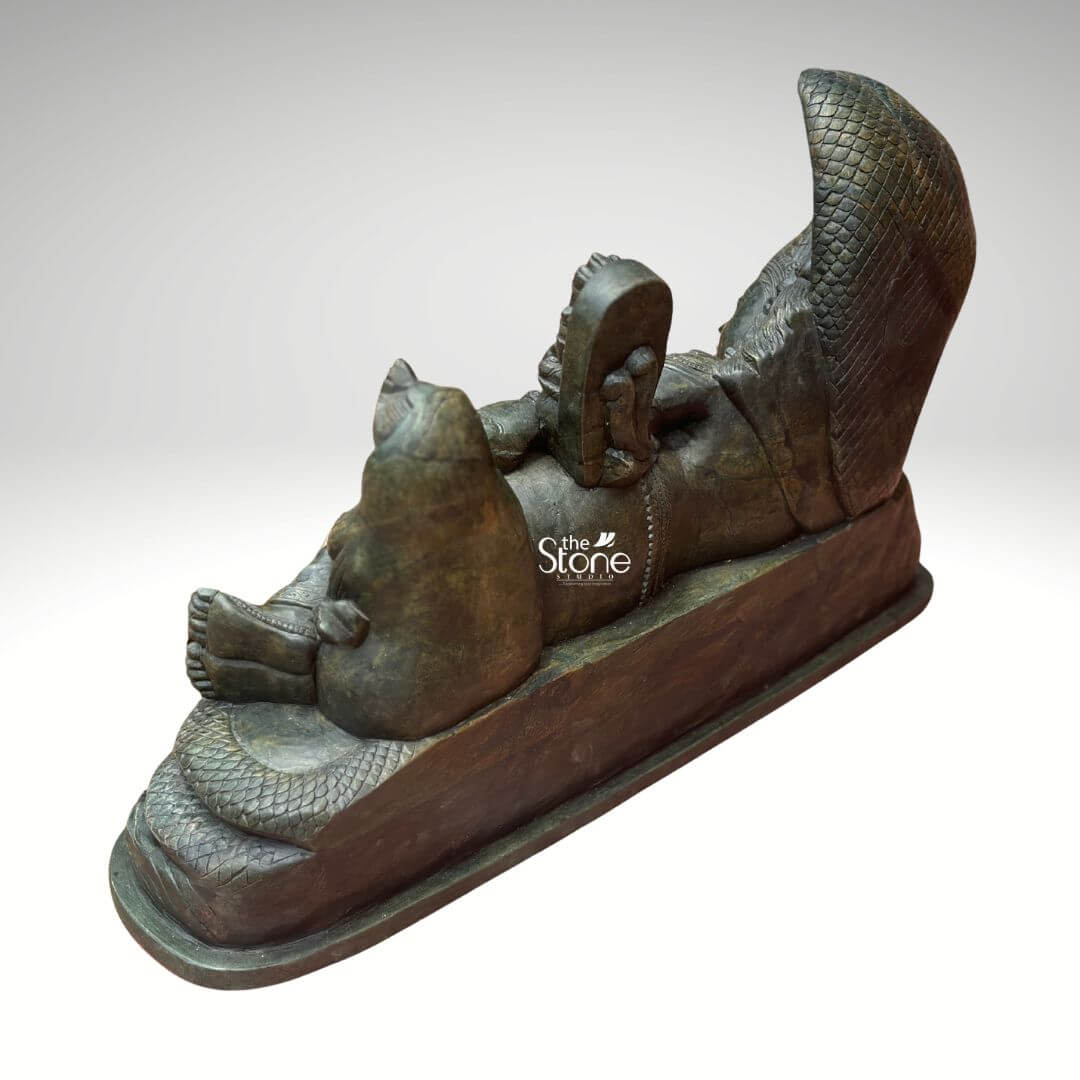 Ananta Shayana Vishnu Statue 20" 9 A striking green stone statue of Sheshashayi Vishnu depicting Lord Vishnu reclining on the cosmic serpent Sheshnag is presented from a rear-right slightly elevated perspective. This angle offers a unique view of the intricate details of the sculpture particularly the coiled body of the serpent and the forms of Lakshmi and Brahma. The deep verdant green of the stone likely soapstone or a similar metamorphic rock lends an ancient and mystical quality to the carving highlighting the profound narrative of cosmic preservation and creation. Lord Vishnu is shown in a state of serene cosmic slumber his form adorned with traditional ornaments though details are partially obscured from this angle. The multi-headed Sheshnag coils intricately beneath him its textured scales and fanned hoods forming a dramatic canopy at Vishnu's head which is visible on the far left. The figure of Goddess Lakshmi is prominently seen seated near Vishnu's feet her back facing the viewer attending to her lord. The small figure of Brahma seated on a lotus emerging from Vishnu's navel is also visible from this perspective illustrating the cyclical emergence of creation. This green stone Sheshashayi Vishnu statue is a powerful and sacred representation of cosmic order preservation and the continuous cycle of time in Hindu mythology. The unique rear-right perspective allows for an appreciation of the full composition and the interplay between the divine figures. The exceptional detail and the choice of green stone often associated with spiritual balance and renewal enhance its profound symbolism making this exquisite statue an impressive and deeply meaningful artifact for a temple home altar.