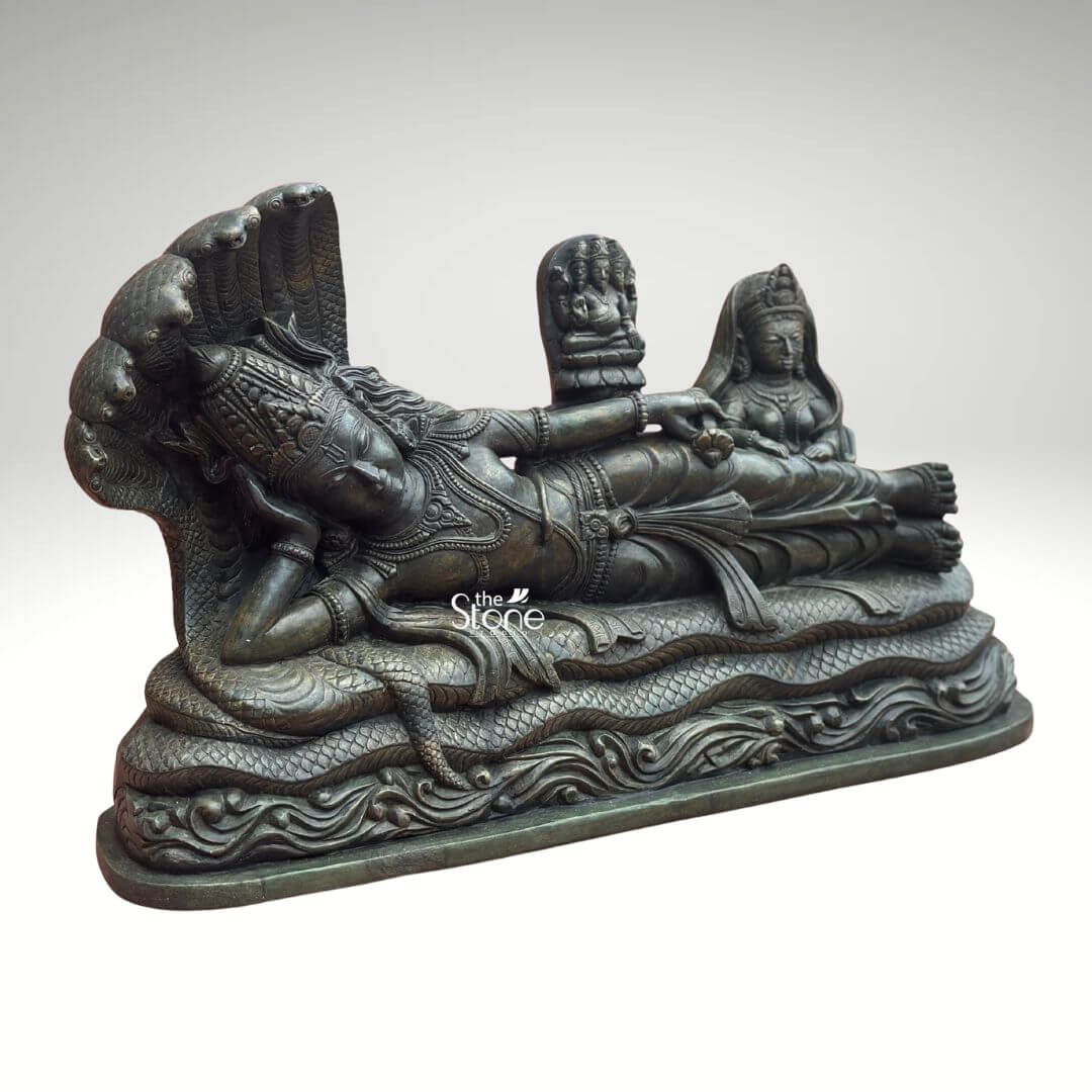 Ananta Shayana Vishnu Statue 20" 10 A majestic green stone statue depicts Lord Vishnu reclining on the cosmic serpent Sheshnag a quintessential representation of cosmic preservation and creation in Hindu mythology. The sculpture viewed from a slightly elevated and oblique angle highlights the intricate details and the profound narrative of Vishnu's Ananta Shayana posture. The deep verdant green of the stone possibly chlorite schist or a similar dark fine-grained material lends an ancient and sacred aesthetic imbuing the artwork with a sense of timelessness and spiritual depth. Lord Vishnu is meticulously carved in a state of serene cosmic slumber his head gently supported by his right arm with an expression of profound peace and detachment from worldly concerns. He is lavishly adorned with traditional Hindu ornaments including an elaborate crown multiple necklac intricate armlets and a gracefully draped dhoti all rendered with exceptional precision. The multi-headed Sheshnag coils beneath him its numerous hoods fanning out to form a grand protective canopy over Vishnu's head with its scales and undulating body intricately rendered to suggest movement and power. From Vishnu's navel a delicate lotus emerges bearing a miniature figure of Brahma the creator god symbolizing the universe's cyclical creation. To Vishnu's right seated attentively is Goddess Lakshmi his divine consort her form equally adorned and sculpted with reverence massaging his feet. This green stone Sheshnag Vishnu statue is a powerful and sacred representation of preservation cosmic order and the cyclical nature of time. The exceptional detail and the choice of green stone often associated with spiritual balance growth and renewal significantly enhance its profound symbolism. This exquisitely crafted statue is an impressive and deeply meaningful artifact ideal for a grand temple a prominent home altar or a spiritual garden inviting contemplation and reverence for the divine cosmic danc and enriching any space with its deep cultural and religious resonance.
