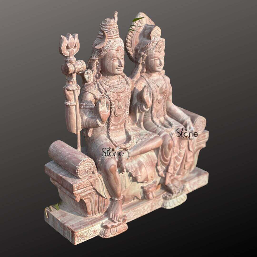 Lord Siva with Maa Parvati Stone Statue 18" 4 An exquisite 18-inch Shiva Parvati Statue meticulously carved from pink stone presented against a dark background as highlighted in an article about such sculptures. The entire piece rendered in a soft warm pinkish-brown hue of natural stone exudes a gentle and serene aesthetic. Lord Shiva and Goddess Parvati are depicted seated side-by-side on an ornate throne. Lord Shiva on the left is depicted with his traditional matted hair adorned with a crescent moon and a small cobra. He holds a trishul in his left hand while his right hand is raised in the Abhaya Mudra. He wears minimal attire and intricate jewelry all carved from the stone. Goddess Parvati sits gracefully to Shiva's right adorned with an elaborate crown and traditional jewelry including necklaces and earrings. Her right hand is also raised in a blessing gesture while her left hand rests elegantly in her lap. Both deities have serene expressions conveying peace and divine harmony. The throne they share is intricately carved with traditional Indian motifs including a lion's face at the front symbolizing strength and royalty. The flowing drapery of their garments is also finely sculpted adding a sense of detail and elegance. A small green leaf is subtly placed behind Parvati's crown. The statue rests on a robust and ornate base also crafted from pink stone featuring lotus petal designs and other decorative patterns. The smooth contours and fine details throughout both figures emphasize a deep sense of tranquility and spiritual harmony. This 18-inch Shiva Parvati statue in its unique pink stone is a powerful representation of devotion and artistic mastery making it an ideal piece for spiritual veneration and artistic appreciation in a home or temple as discussed in the accompanying article.